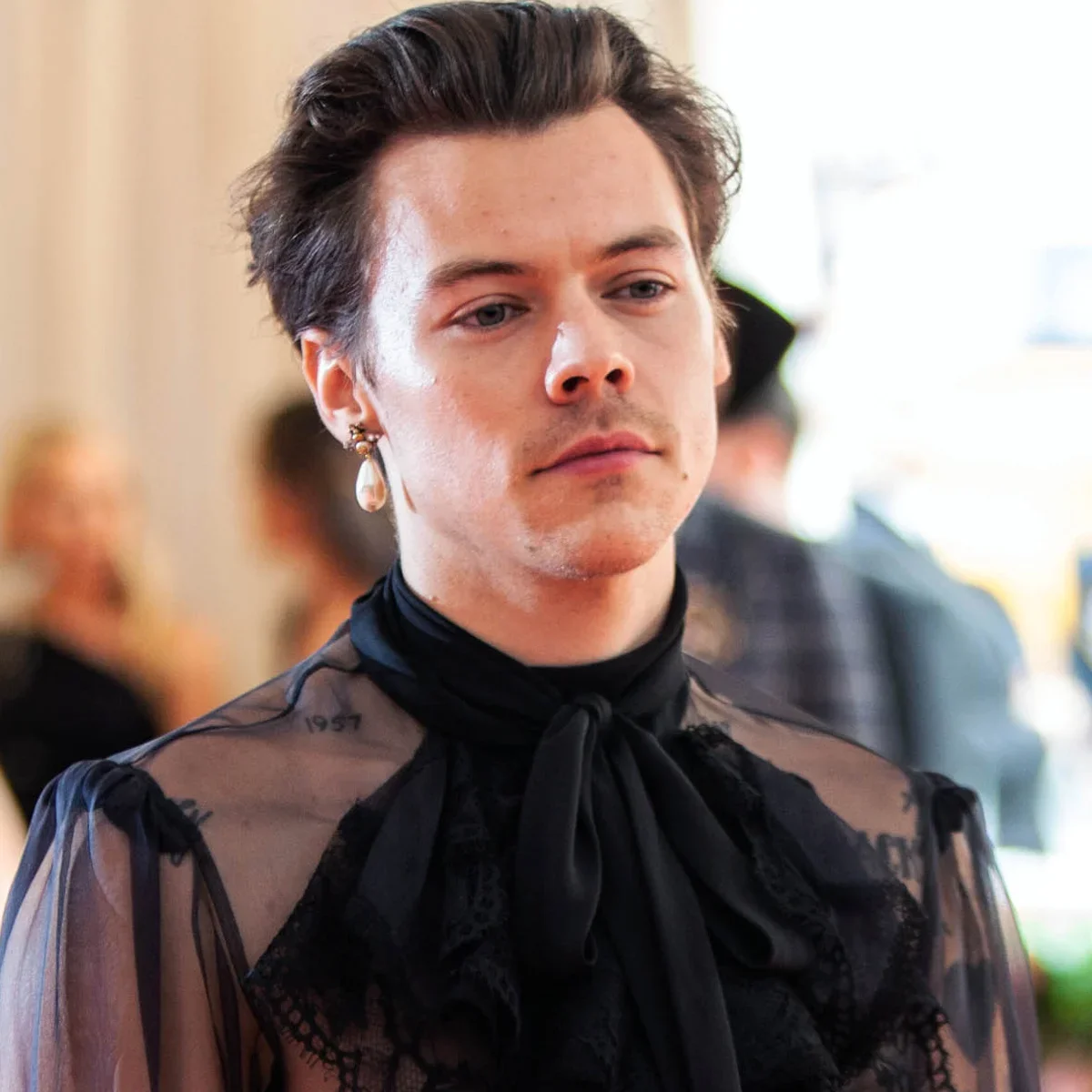 Young man with dark hair, wearing a sheer black lace top with a bow tie, and a pearl earring, at an indoor event.