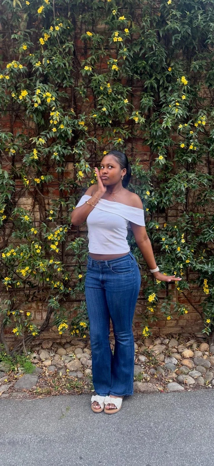 A young woman in a white off-the-shoulder top and blue jeans stands in front of a brick wall covered in yellow-flowered green climbing plants, posing with her hand on her face.
