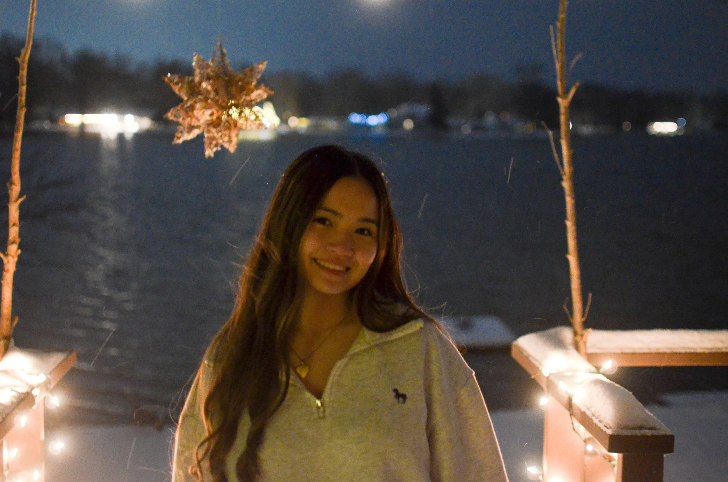 A woman with long brown hair smiling outdoors at night, standing on a snowy pathway decorated with string lights, with a blurred background of snow and distant lights.