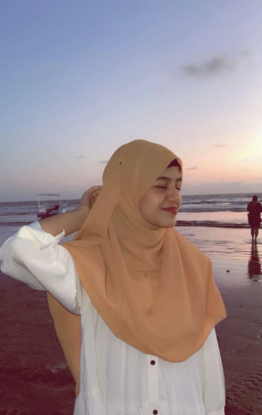 A woman wearing a beige hijab and white dress smiling with her eyes closed on a beach at sunset, with the ocean and a boat in the background.
