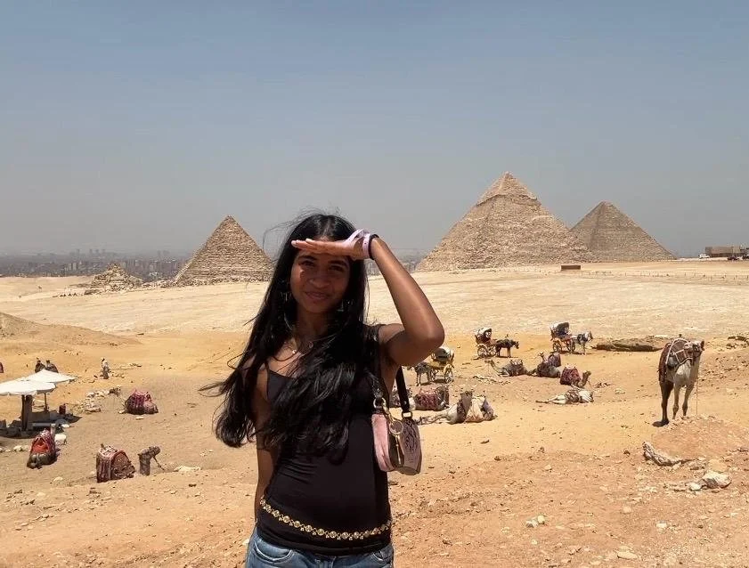 A woman with long black hair shielding her eyes with her hand stands in front of the pyramids at Giza, Egypt, with camels and touristic activity in the desert.