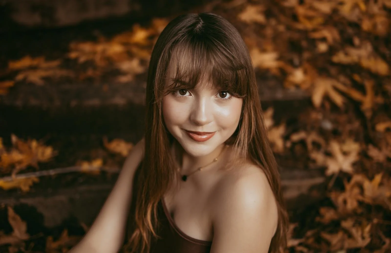 A young woman with long, brown hair and bangs, smiling and looking at the camera, with autumn leaves in the background.