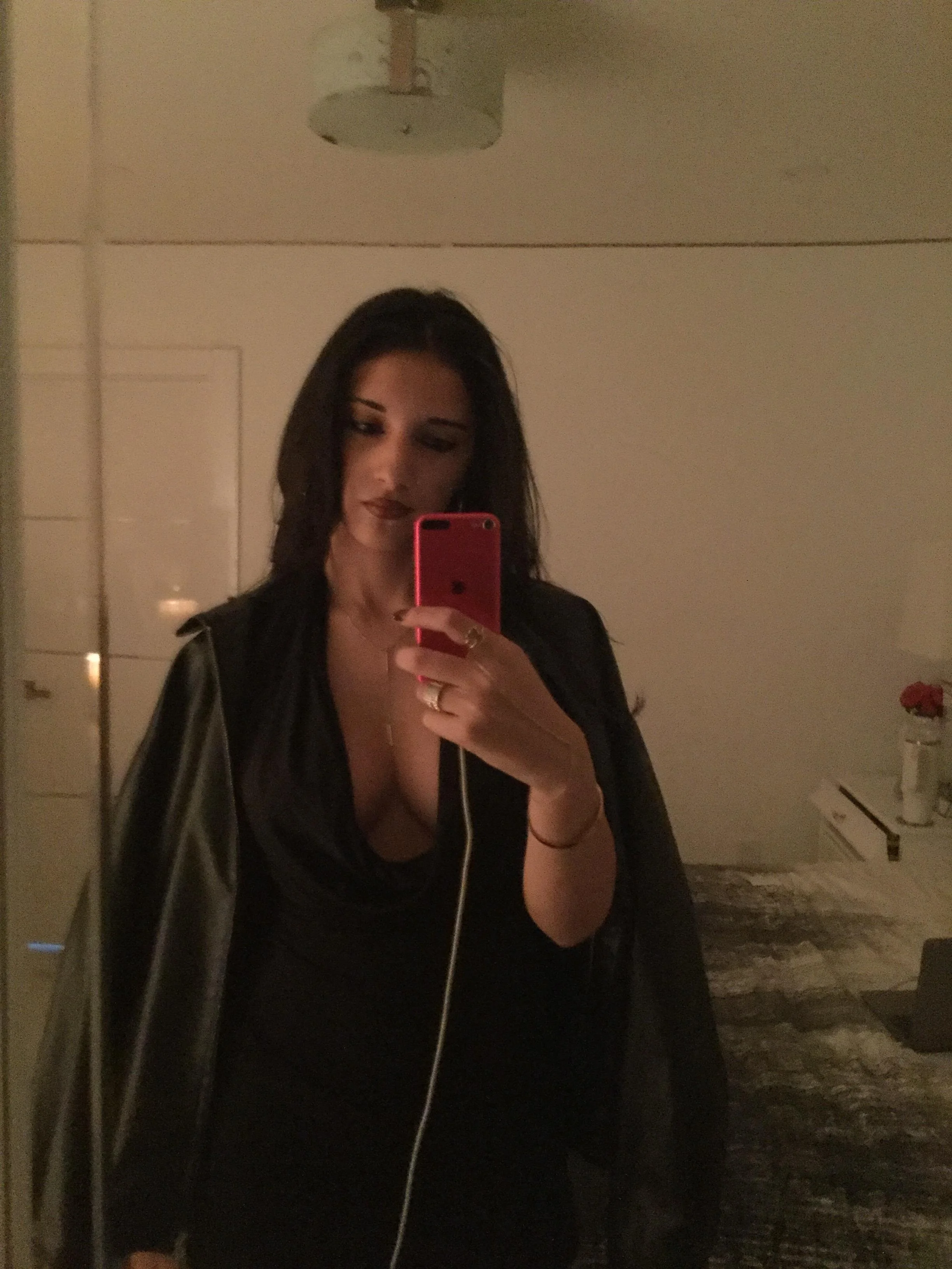 A woman taking a mirror selfie with a pink phone in a dimly lit room, wearing dark clothing and a leather jacket.