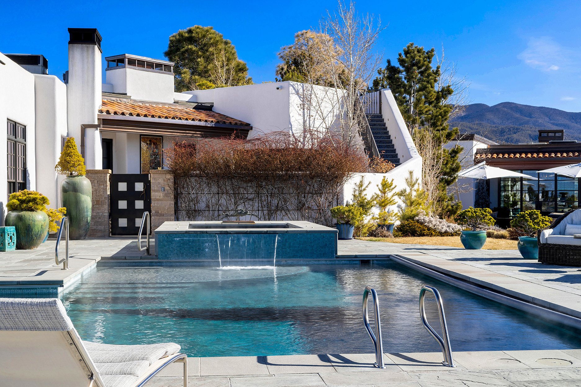 Luxury outdoor area with swimming pool, hot tub, and modern architecture. Features large planters, patio furniture, and a scenic mountain backdrop.