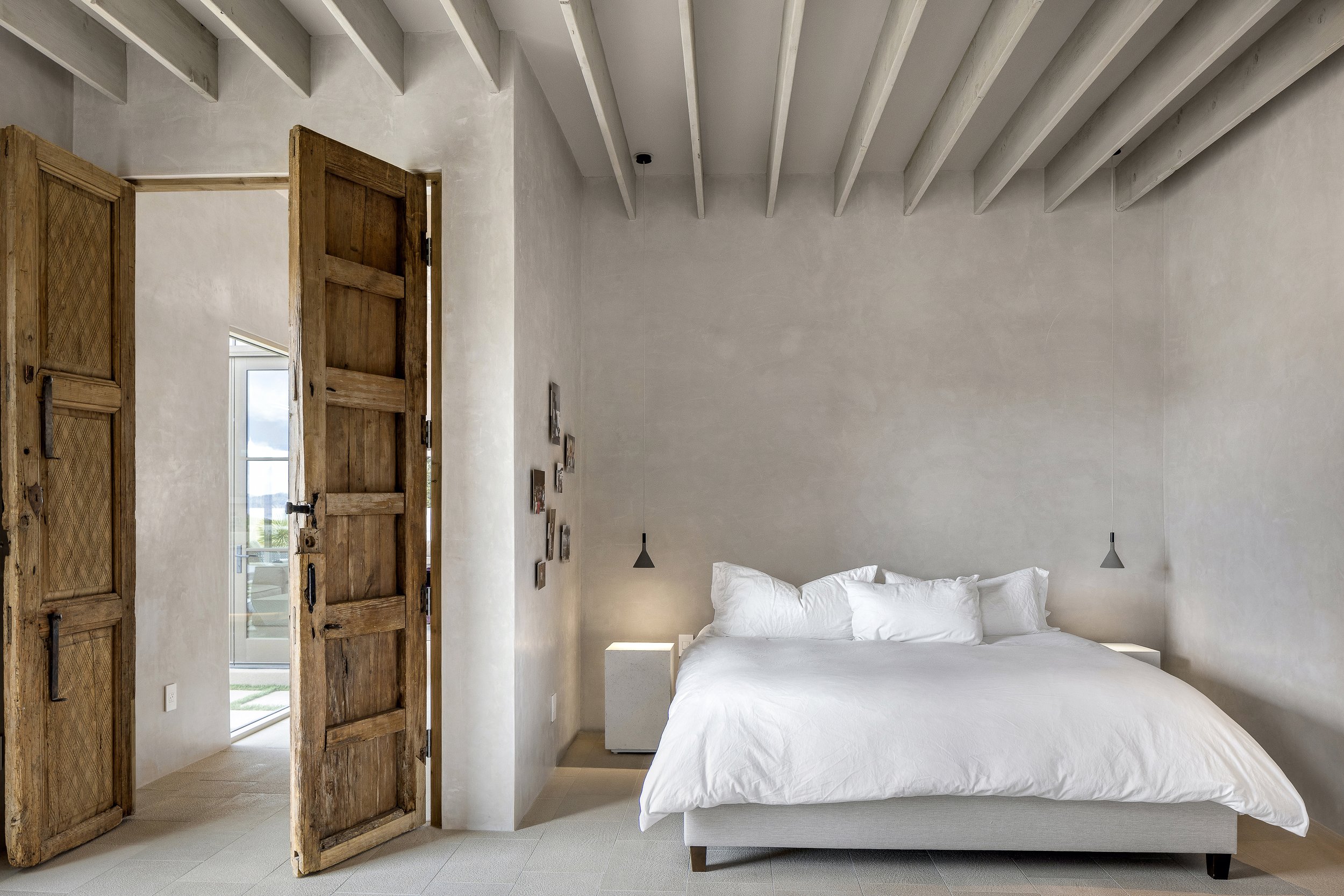 Minimalist bedroom with wooden double doors, a bed with white bedding, concrete walls, pendant lights, and exposed ceiling beams.