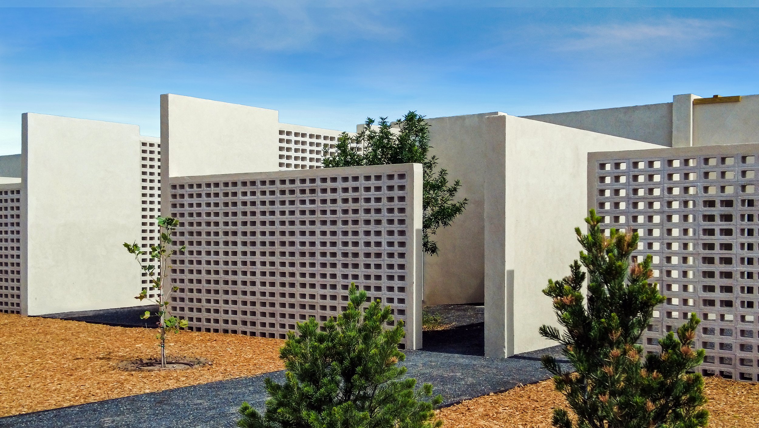 Outdoor area with modern breezeblock walls, small trees, mulch ground cover, and a clear blue sky.