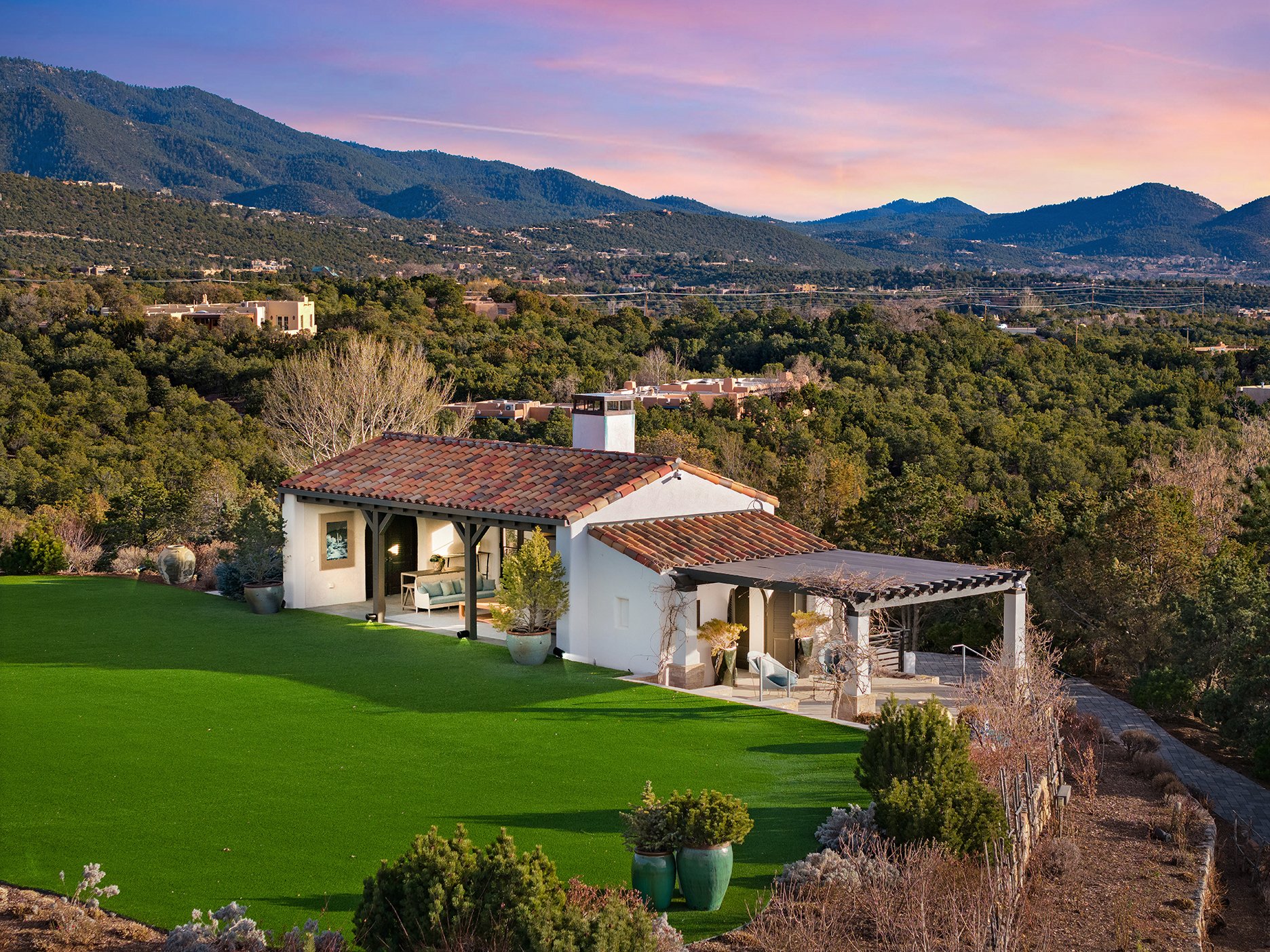 A small white house with a terracotta tile roof situated within a large green lawn and surrounded by trees, with mountains and a vibrant sky in the background.