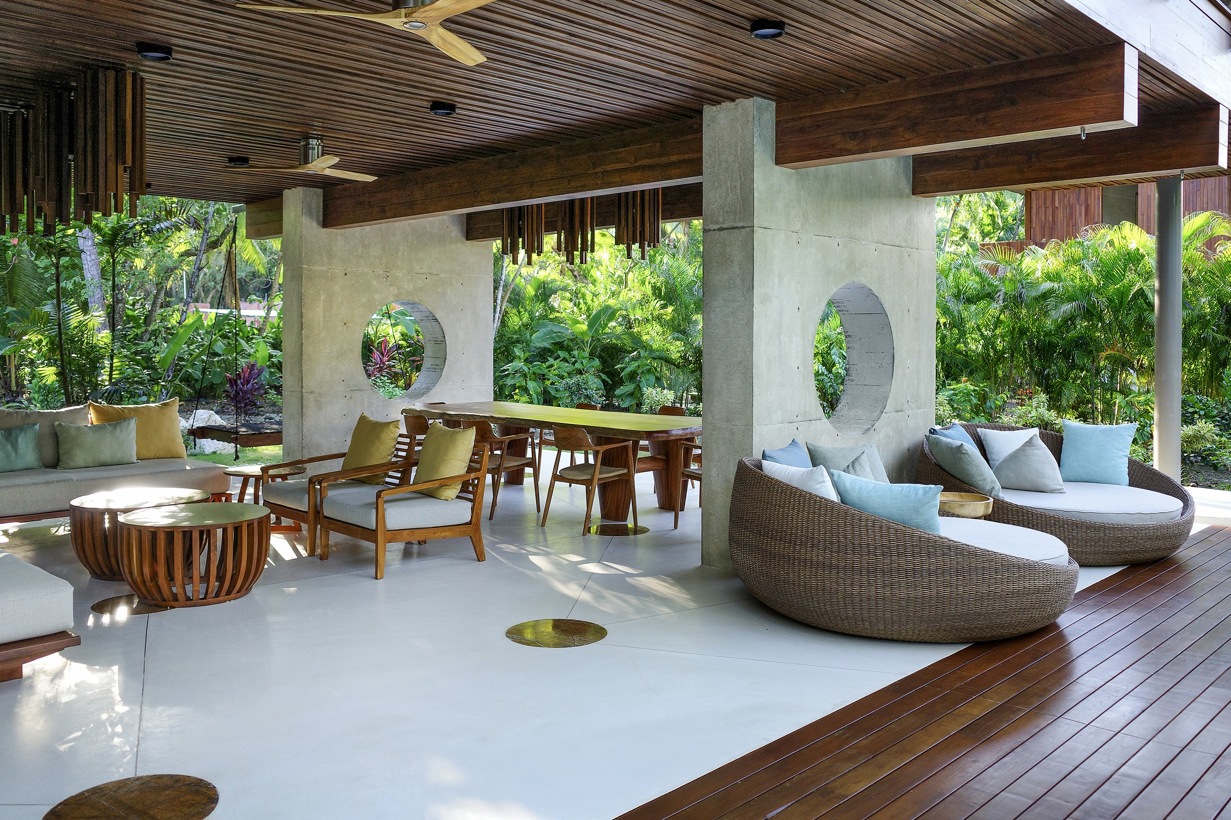 Modern outdoor patio with wooden ceiling and furniture, concrete pillars with circular cutouts, cushioned seating, round tables, and lush green garden surrounding.