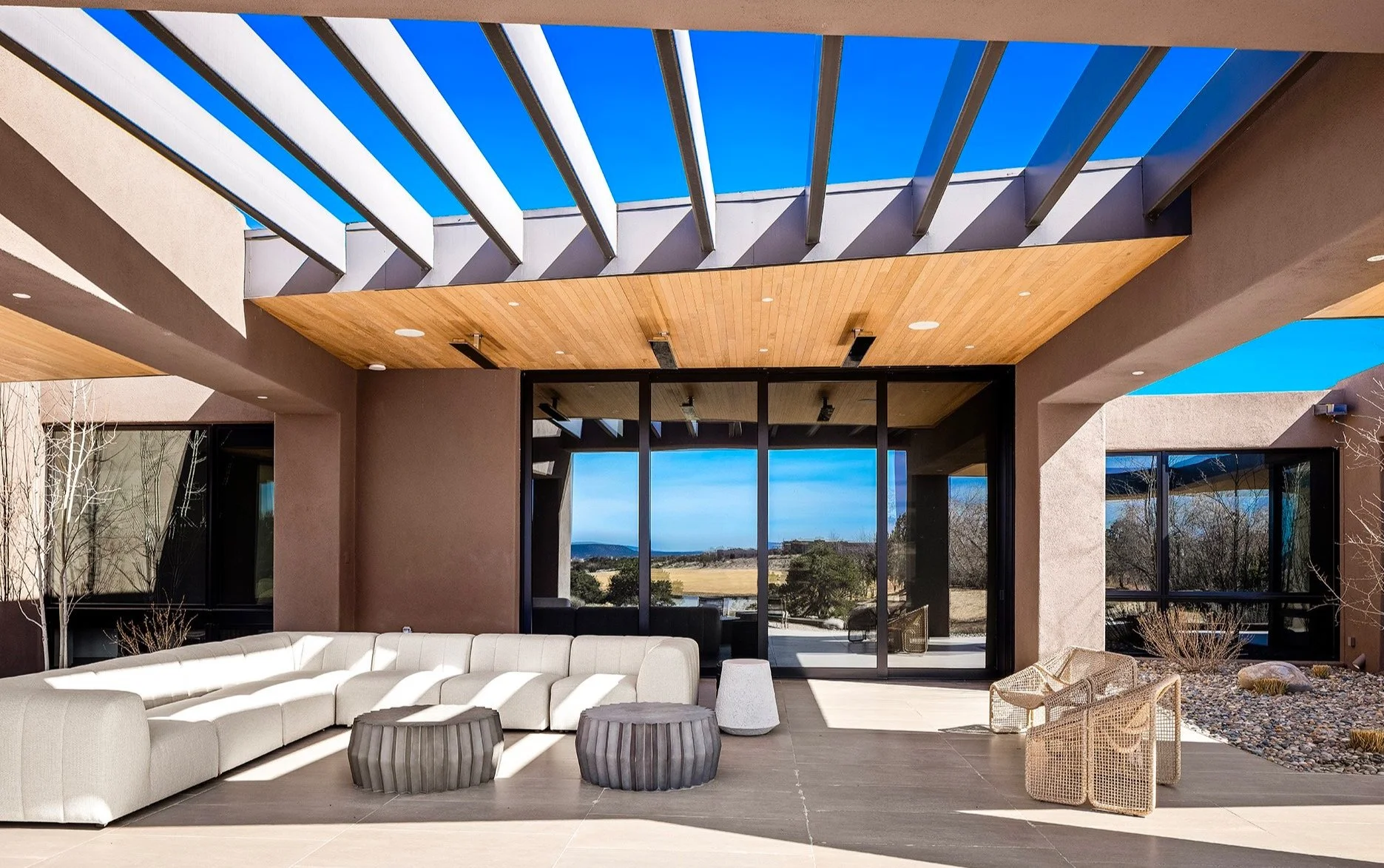 Modern patio with white L-shaped sofa, two round tables, wicker chair set, partial glass roof, and view of outdoor landscape.