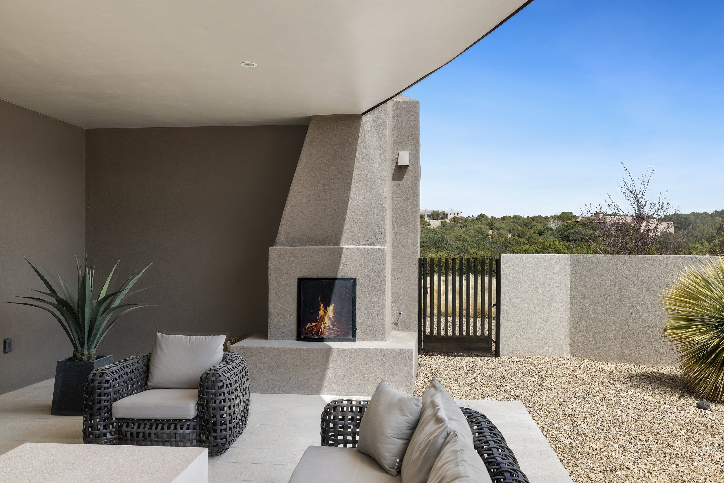 Outdoor patio area with a modern fireplace, gray seating, a potted plant, and a view of a gravel yard and distant greenery.