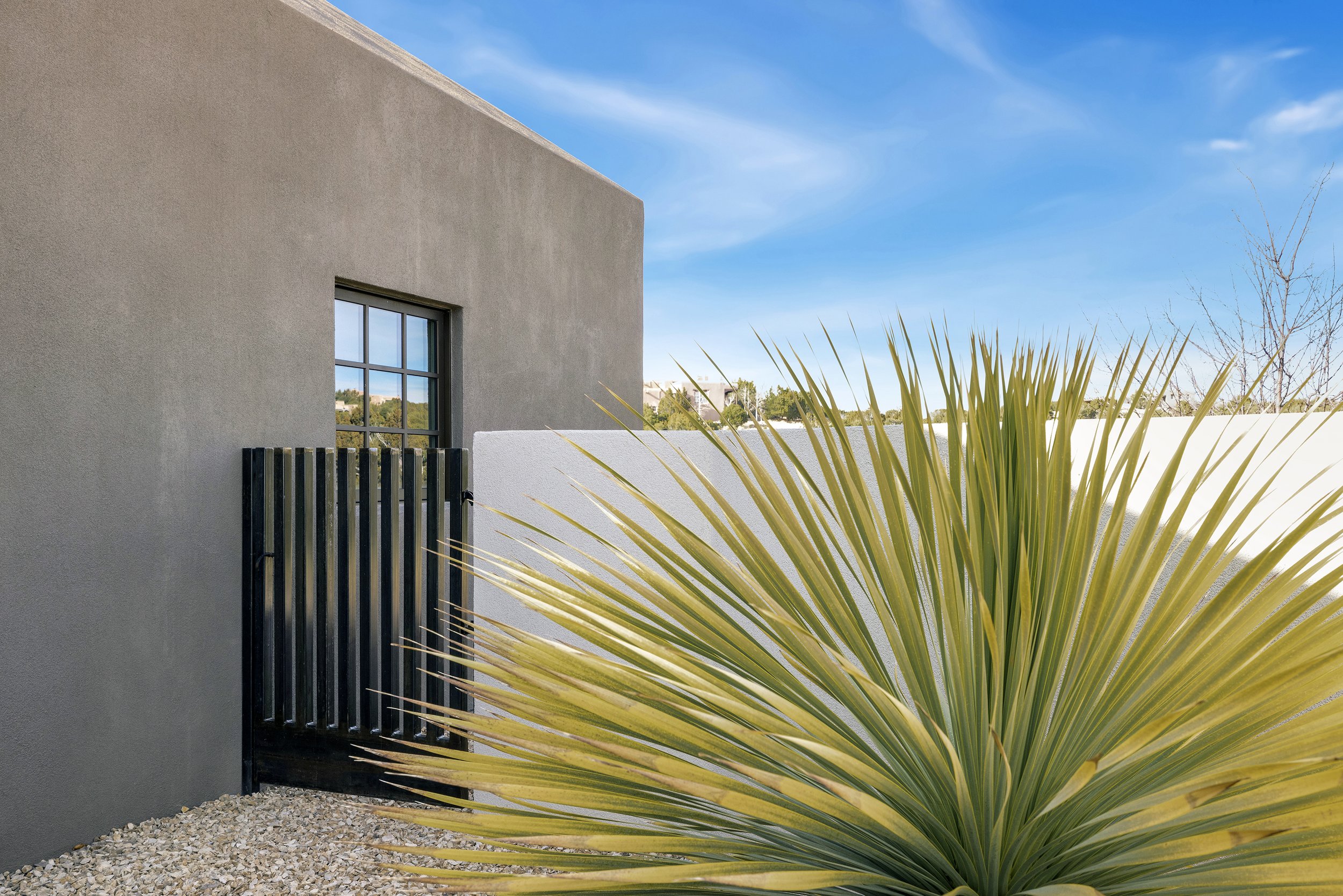 Modern gray stucco building with a black gate, gravel foreground, and spiky desert plant under a blue sky.