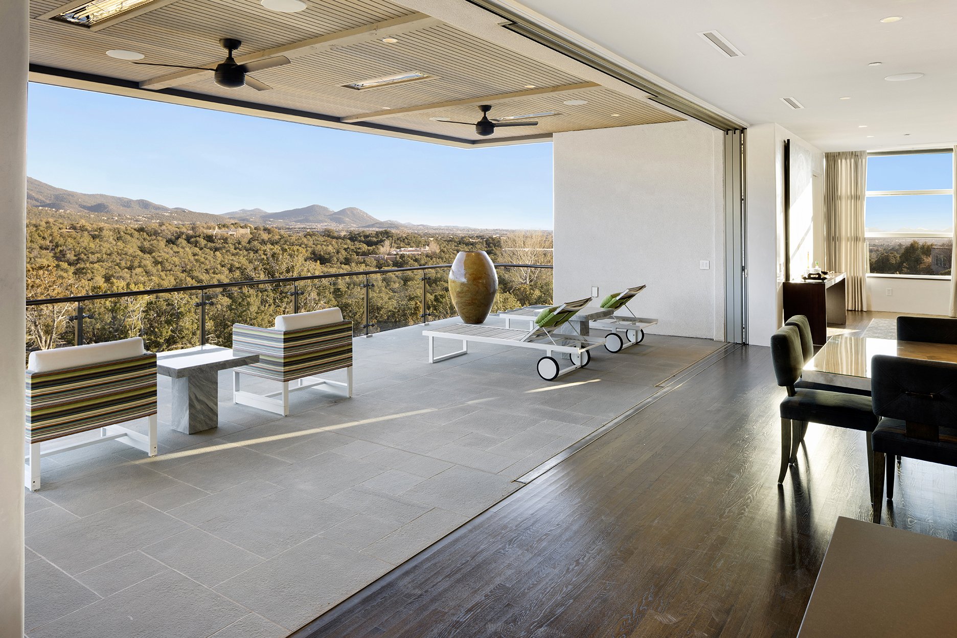 Modern living room with large open balcony, featuring comfortable chairs, a table, and deck chairs overlooking a scenic view of trees and mountains under a clear sky.