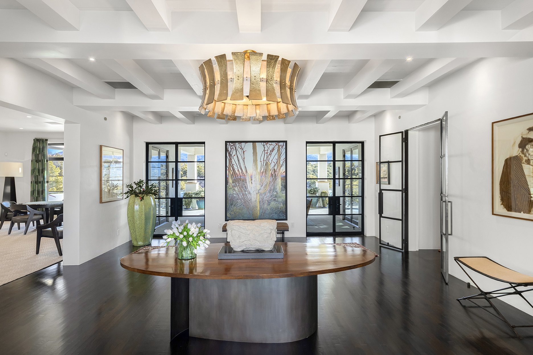 Elegant living room with large chandelier, wooden table, and art pieces. French doors lead to a patio. Modern decor with dark flooring and white walls.