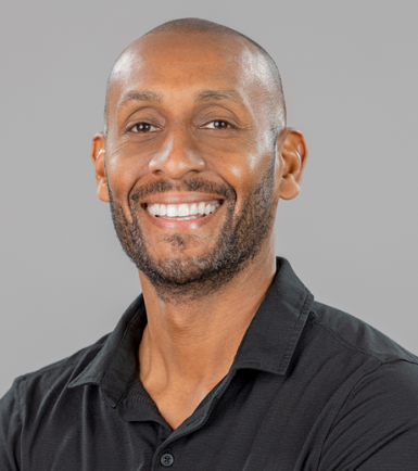 A smiling man with a bald head and a beard, wearing a black collared shirt, against a gray background.