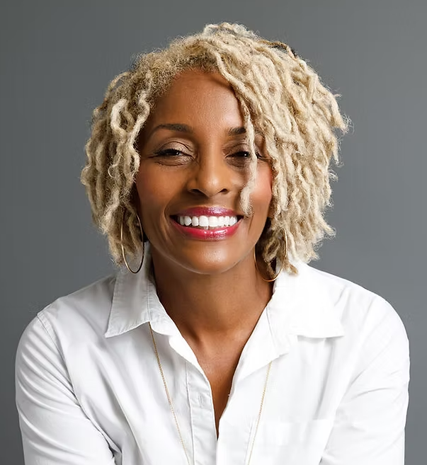 Smiling woman with blonde, curly dreadlocks wearing a white shirt and hoop earrings, against a gray background.