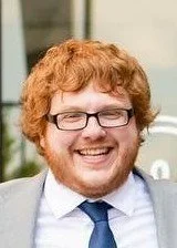 Red headed male with glasses, beard, and mustached.  He is smiling while wearing a gray suit, white button shirt, and blue tie.