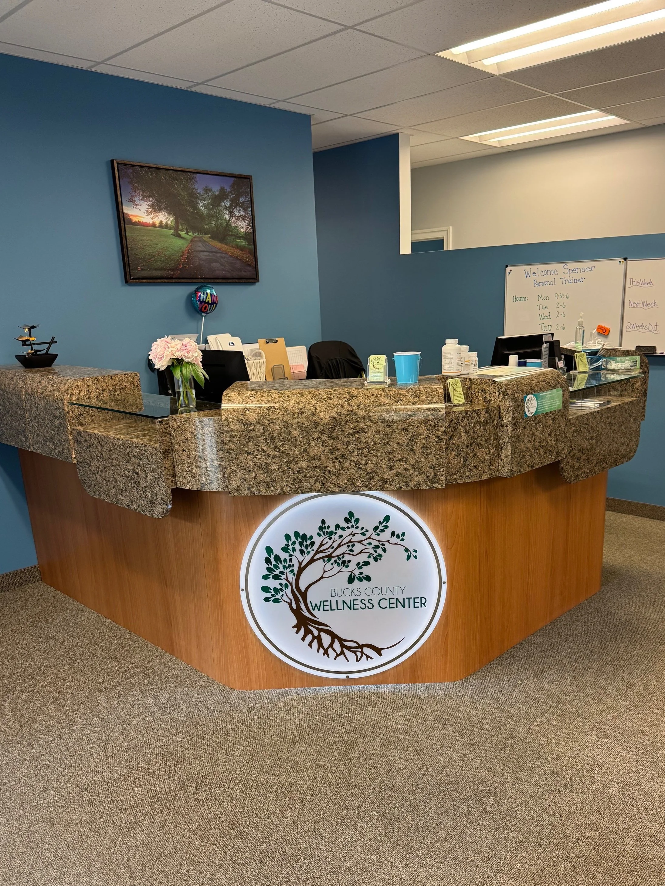 Reception desk at Bucks County Wellness Center with a granite top and a wooden front, featuring a circular logo with a tree. Chiropractic, Massage,  Personal Training and Yoga Services in New Britain, PA near Doylestown and Chalfont.