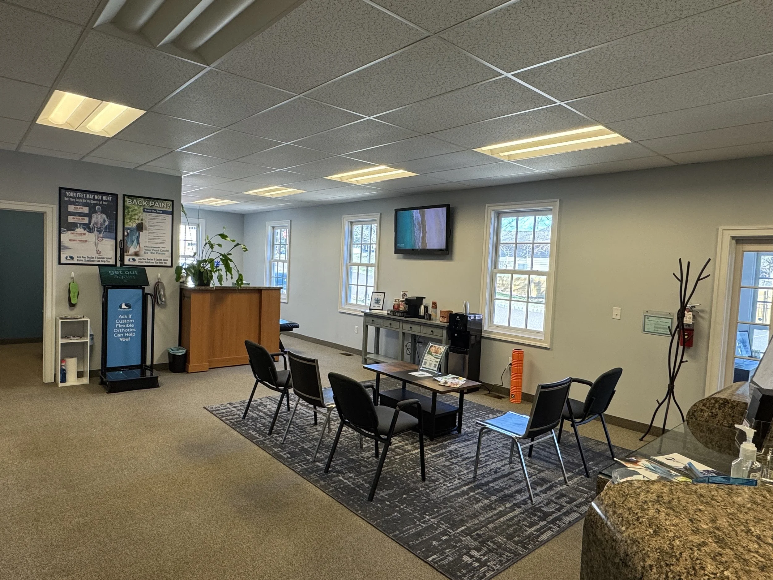 Waiting room with chairs, a coffee station, promotional posters, a wall-mounted TV, and a reception desk. Chiropractor and massage located in the Doylestown, New Britain, and Chalfont area.