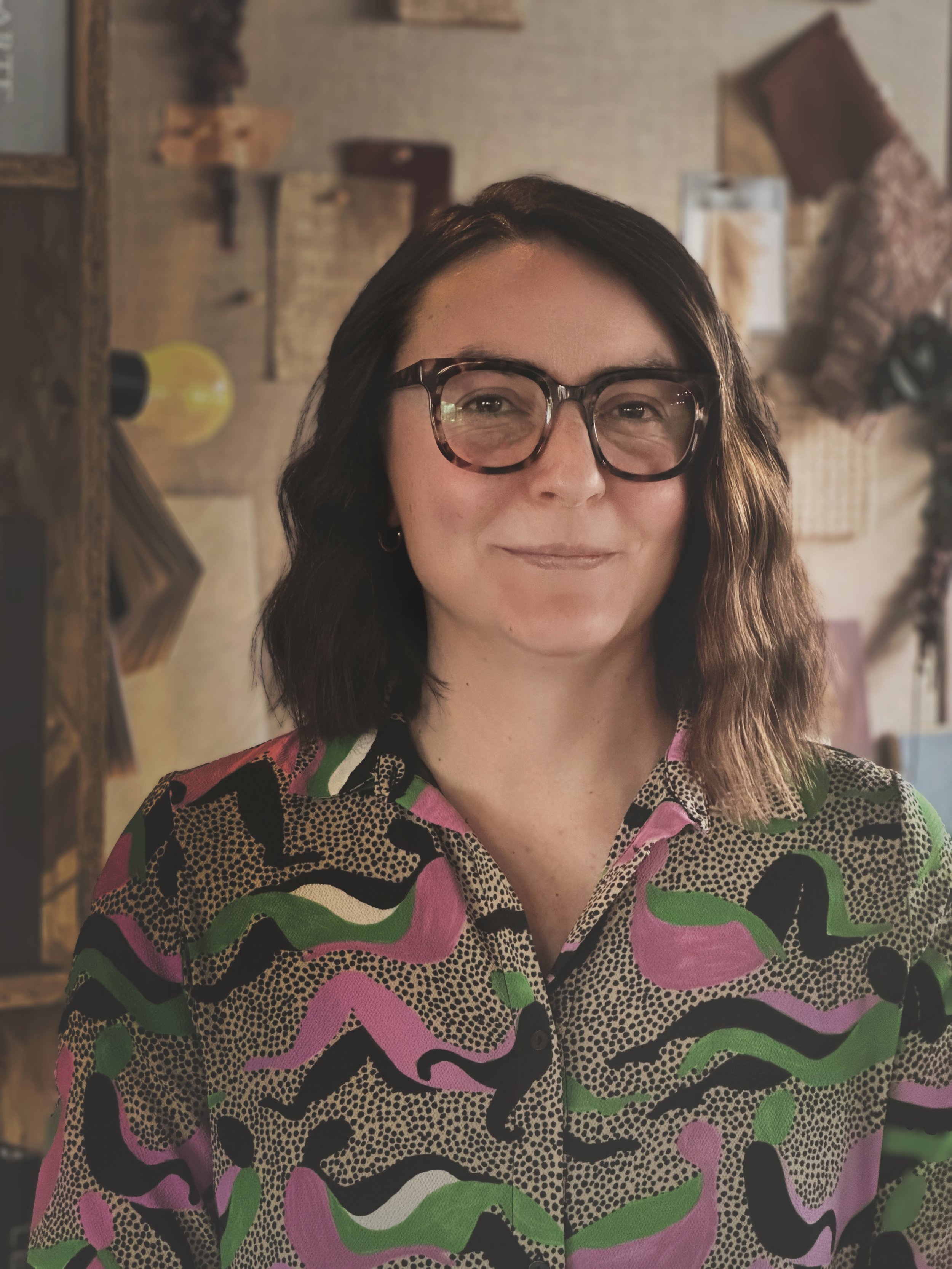 A confident female interior designer wearing a brightly coloured patterned blouse, stood in front of a mood board in a top interior design studio.