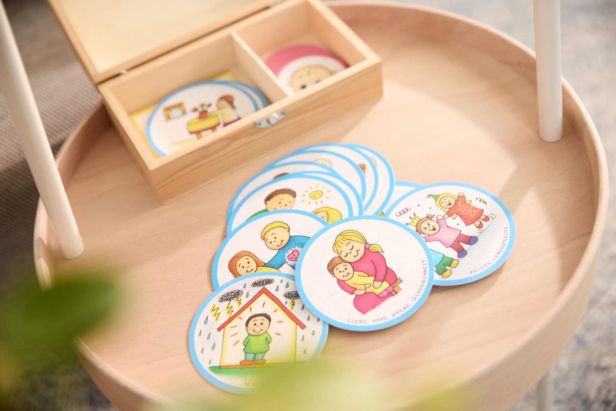 Runde Karten mit bunten Illustrationen, die verschiedene emotionale Ausdrücke und Situationen von Kindern darstellen, liegen auf einem runden Holztisch.