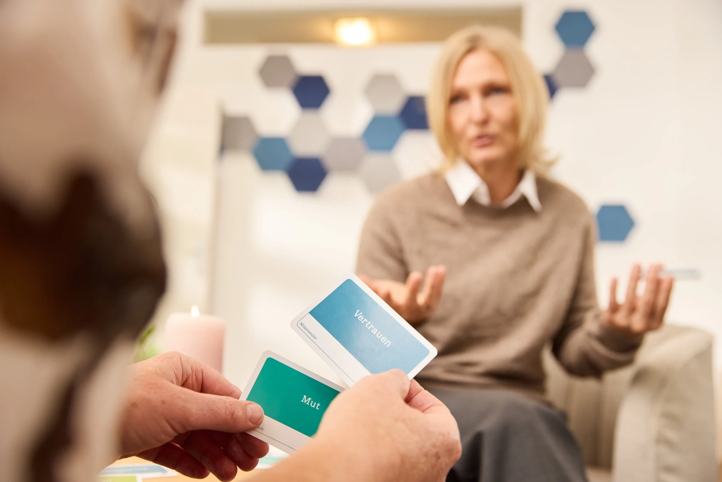 Zwei Personen sitzen sich gegenüber, eine Person hält Karten mit Begriffen wie Vertrauen und Mut. Im Hintergrund eine Unternehmensdeko mit blauen und grauen Hexagonen.