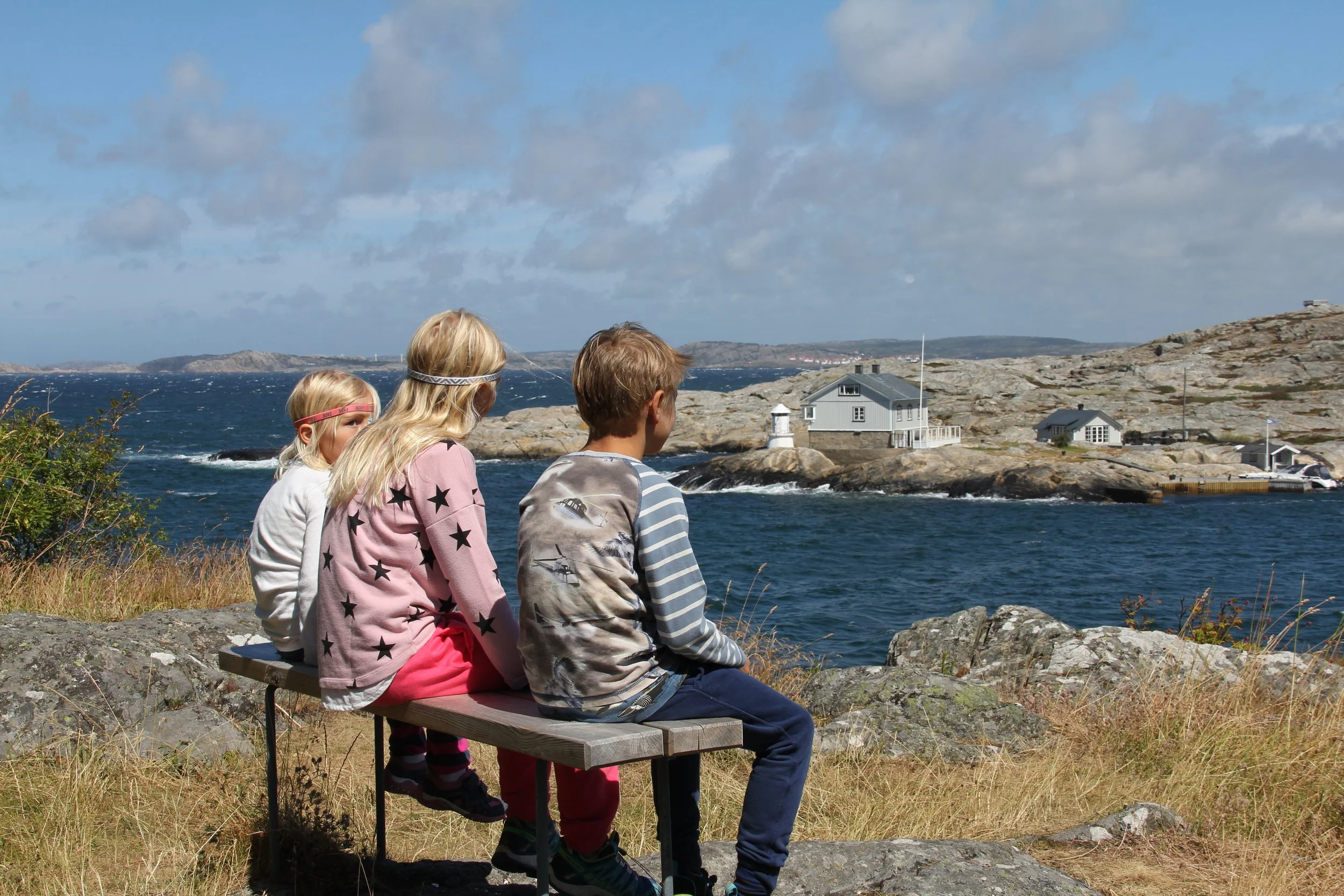 Drei Kinder sitzen auf einer Bank an der Küste und schauen auf das Wasser und eine kleine Insel mit Häusern und Leuchtturm.