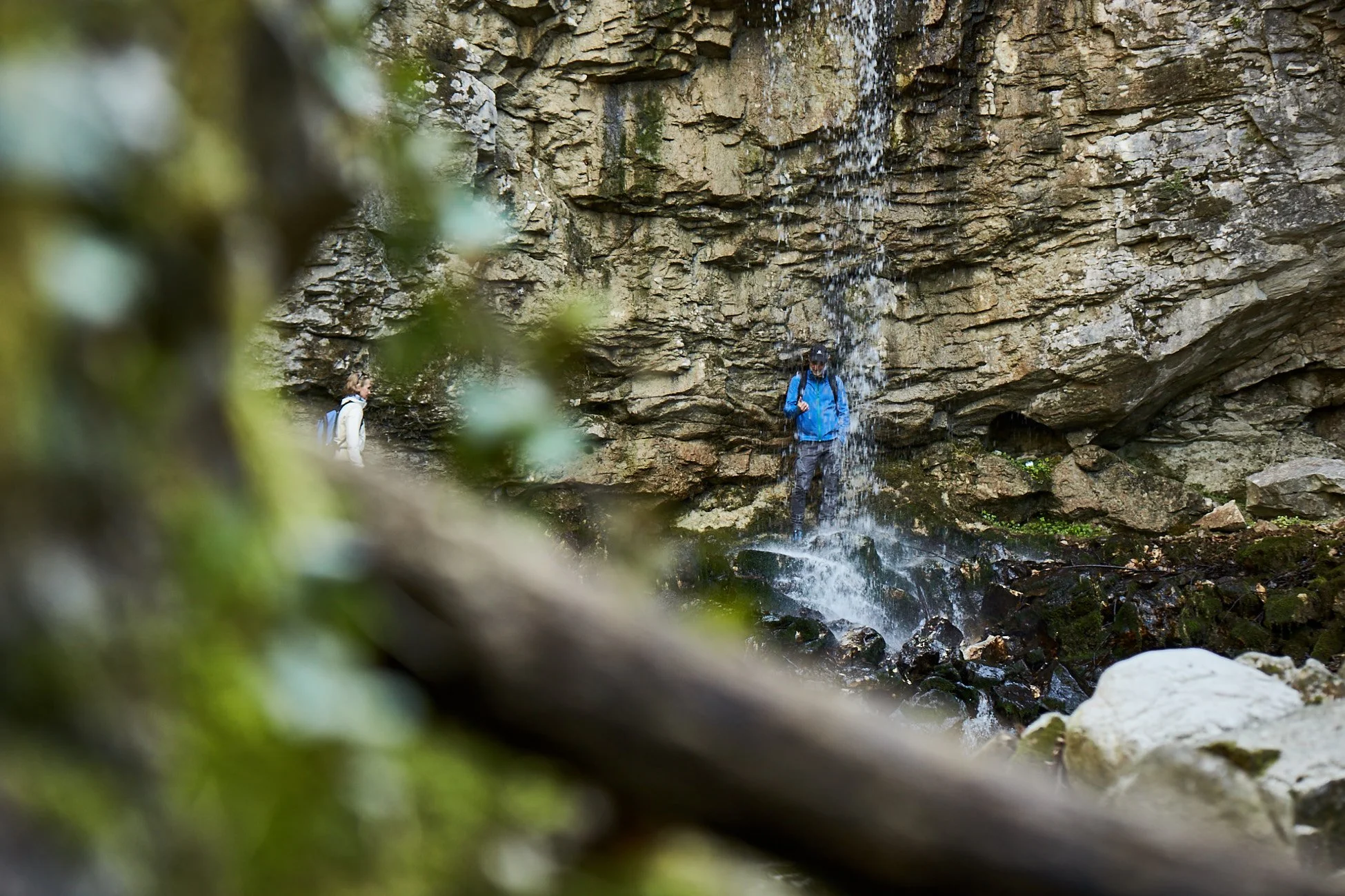 Zwei Wanderer stehen vor einem kleinen Wasserfall in einer felsigen Schlucht, umgeben von Natur.