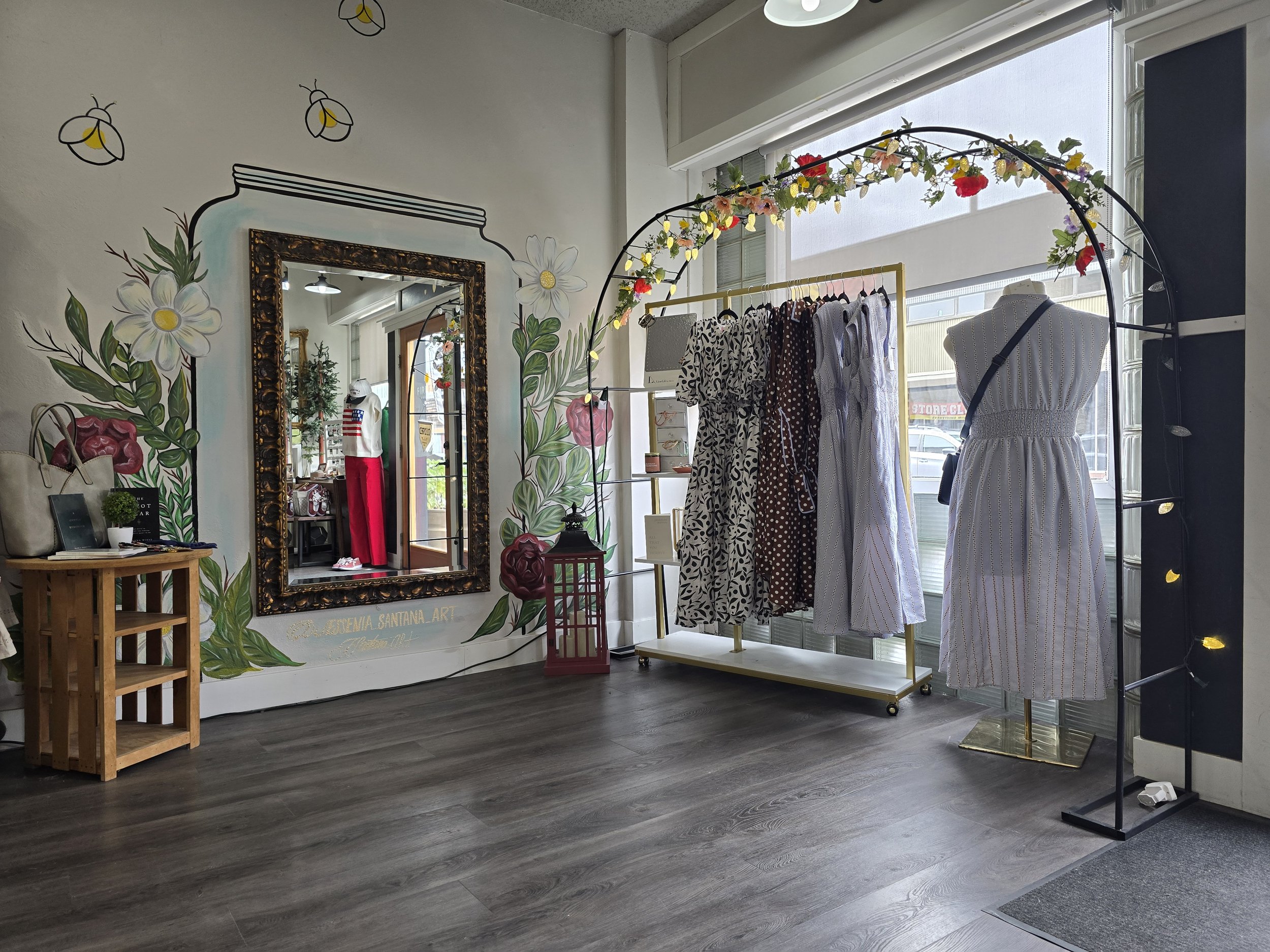 A boutique clothing store with a wall decorated with painted flowers and a large mirror, displaying a mannequin dressed in a striped dress and a clothing rack with patterned dresses, illuminated by natural light from large windows.