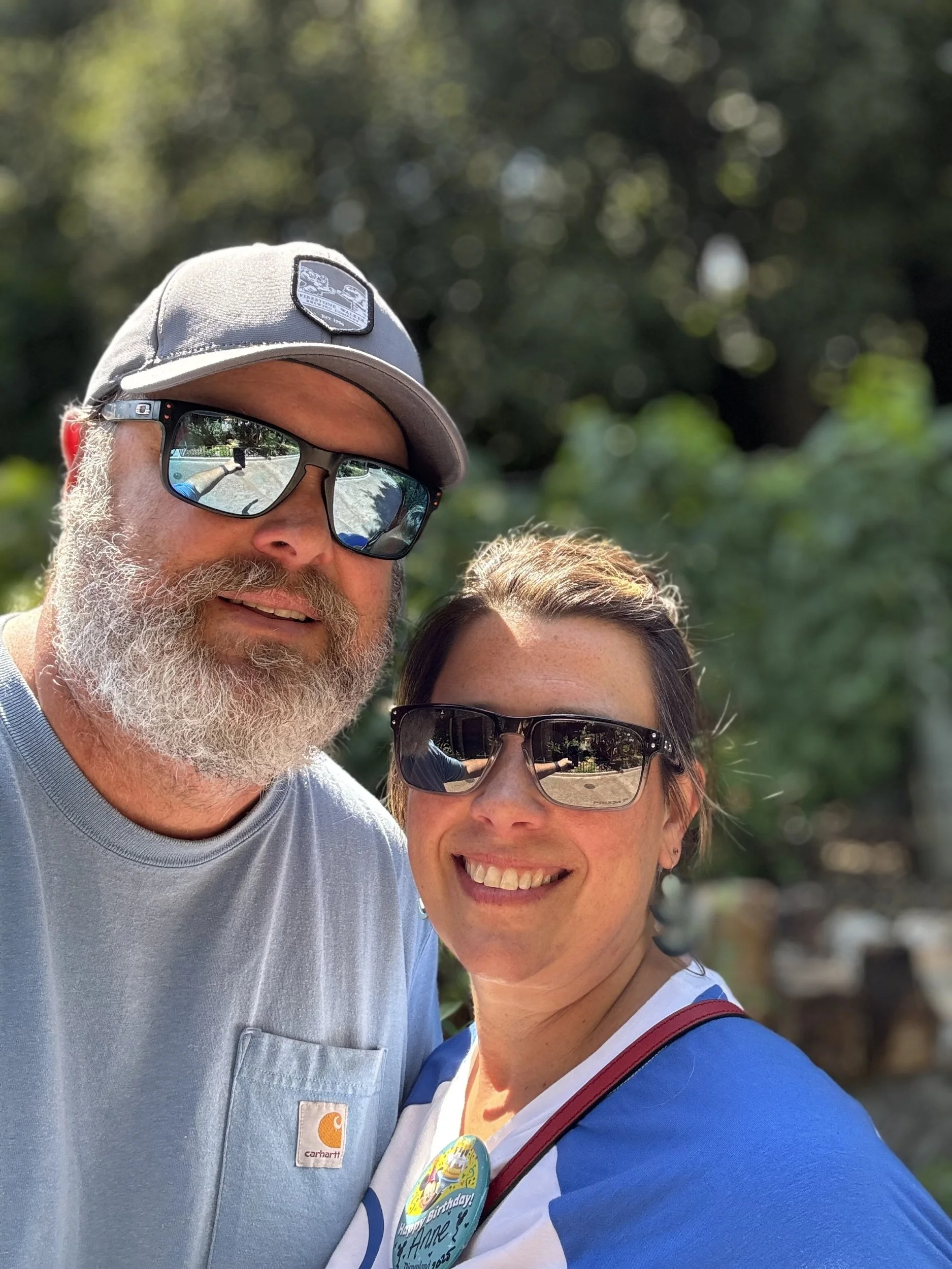 A man with a gray beard wearing sunglasses and a gray Carhartt shirt, with a woman wearing sunglasses and a blue and white shirt, posing outdoors with green trees in the background.