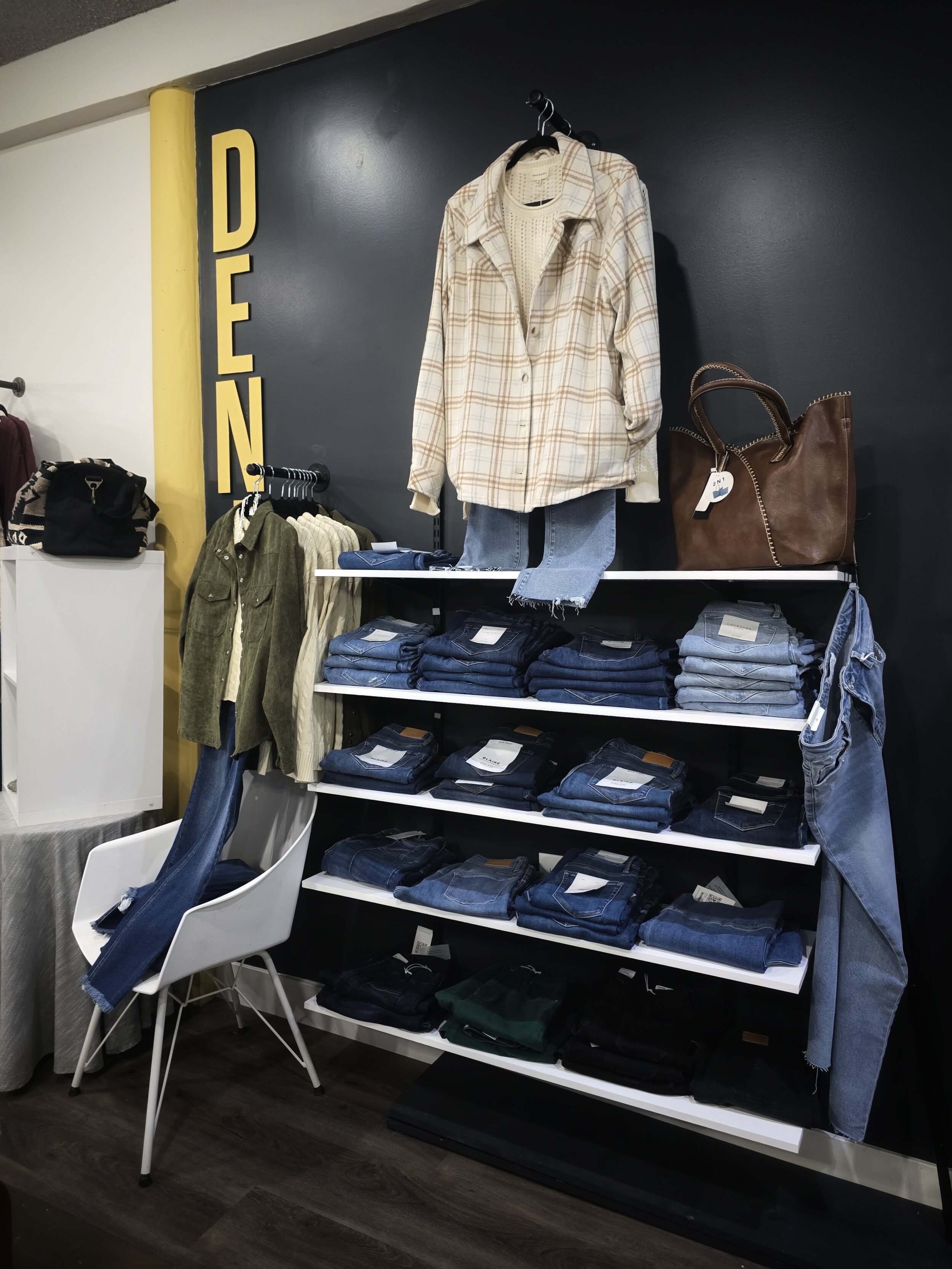 Display of men's clothing with a sign reading 'DENIM', including a plaid shirt, jeans, handbags, and a chair with jeans draped over it in a retail store.