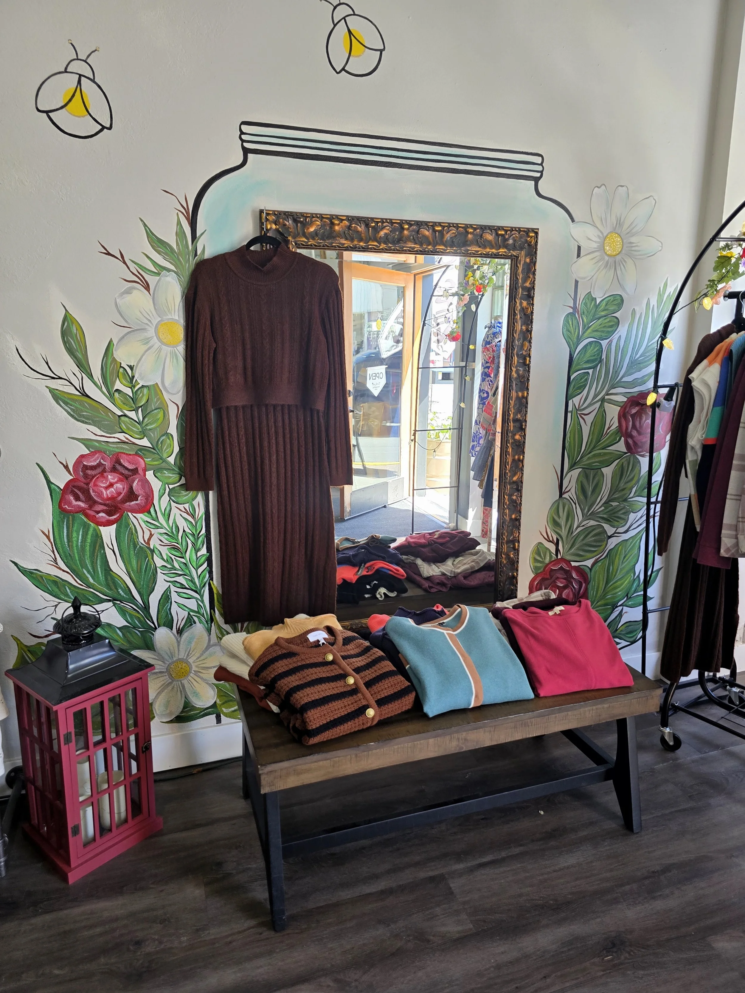 A retail store display featuring a brown knitted dress hanging on the wall, surrounded by colorful floral wall art. A wooden table below holds folded clothes in various colors, and a pink lantern is placed on a small black table to the left of the di