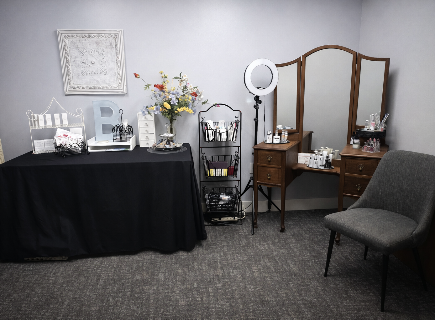 A makeup station with a large mirror, a table with skincare and beauty products, a ring light, and a gray armchair in a room with gray walls and carpet.