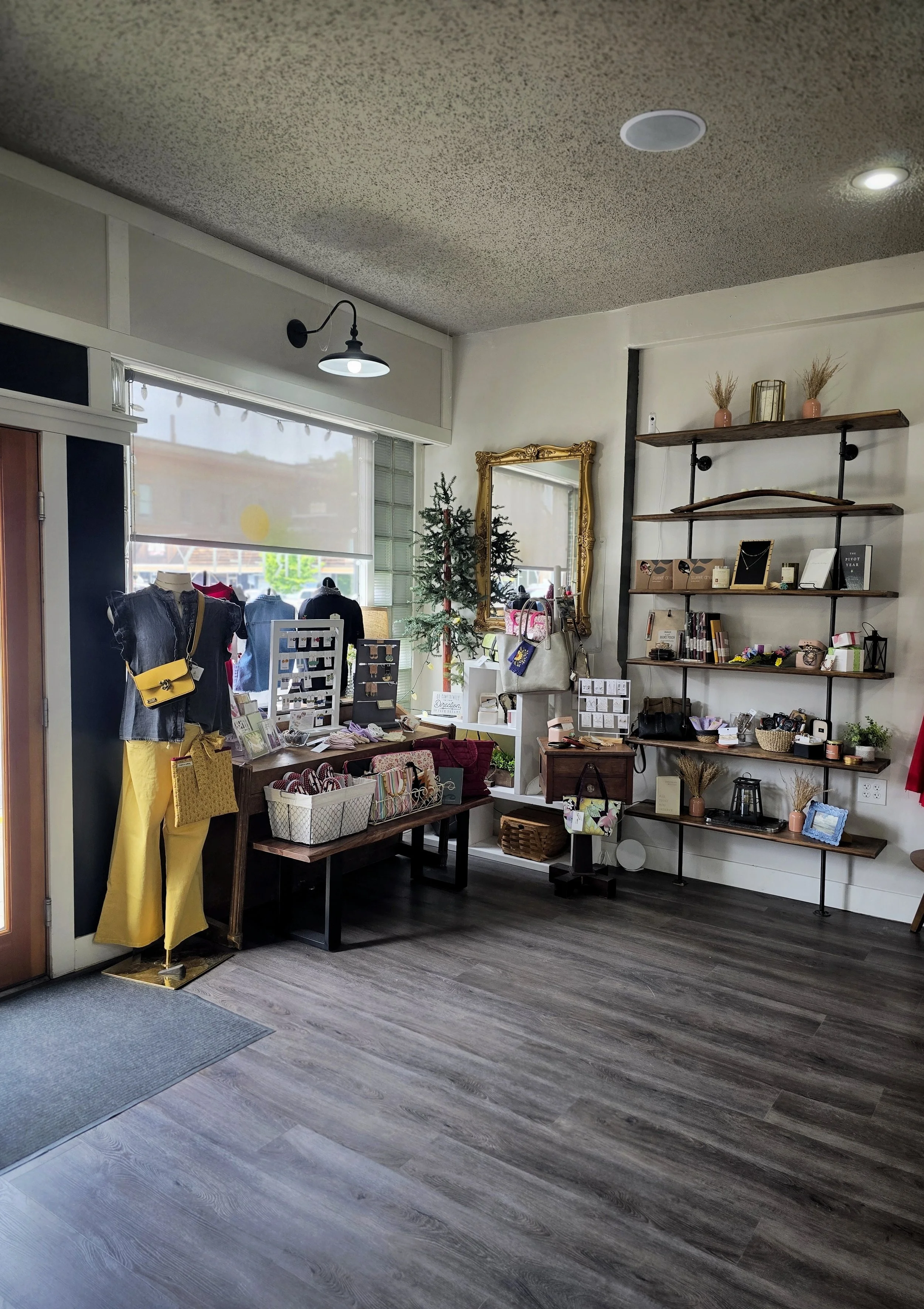 Interior of a small boutique or gift shop with wooden and metal shelves displaying decorative items, jewelry, and bags. Mannequins dressed in clothing near the window, which has a tree and a gold mirror. The floor is wooden, and there is a mat at the