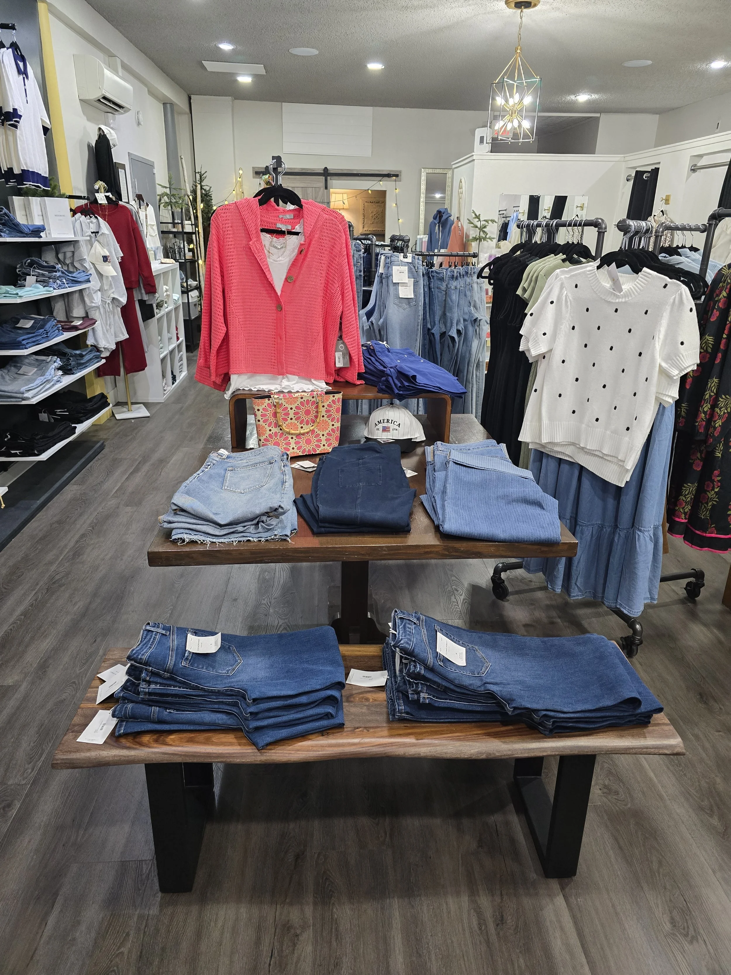 Clothing store with racks of jeans, sweaters, and shirts, including a pink cardigan, white polka-dot sweater, and various denim jeans on tables.