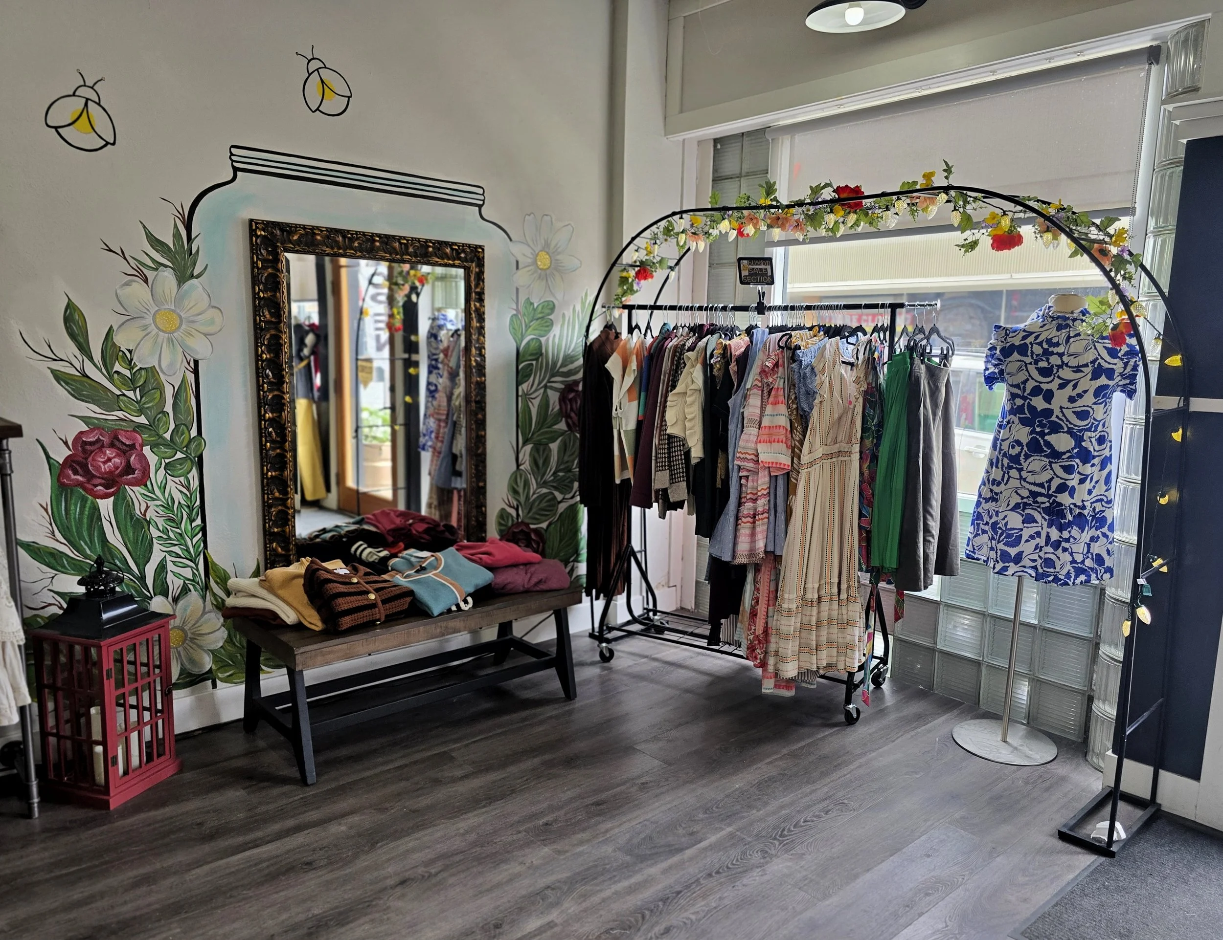Clothing store display with colorful dresses on a rack, a mirror, and wall art with flowers and fireflies.