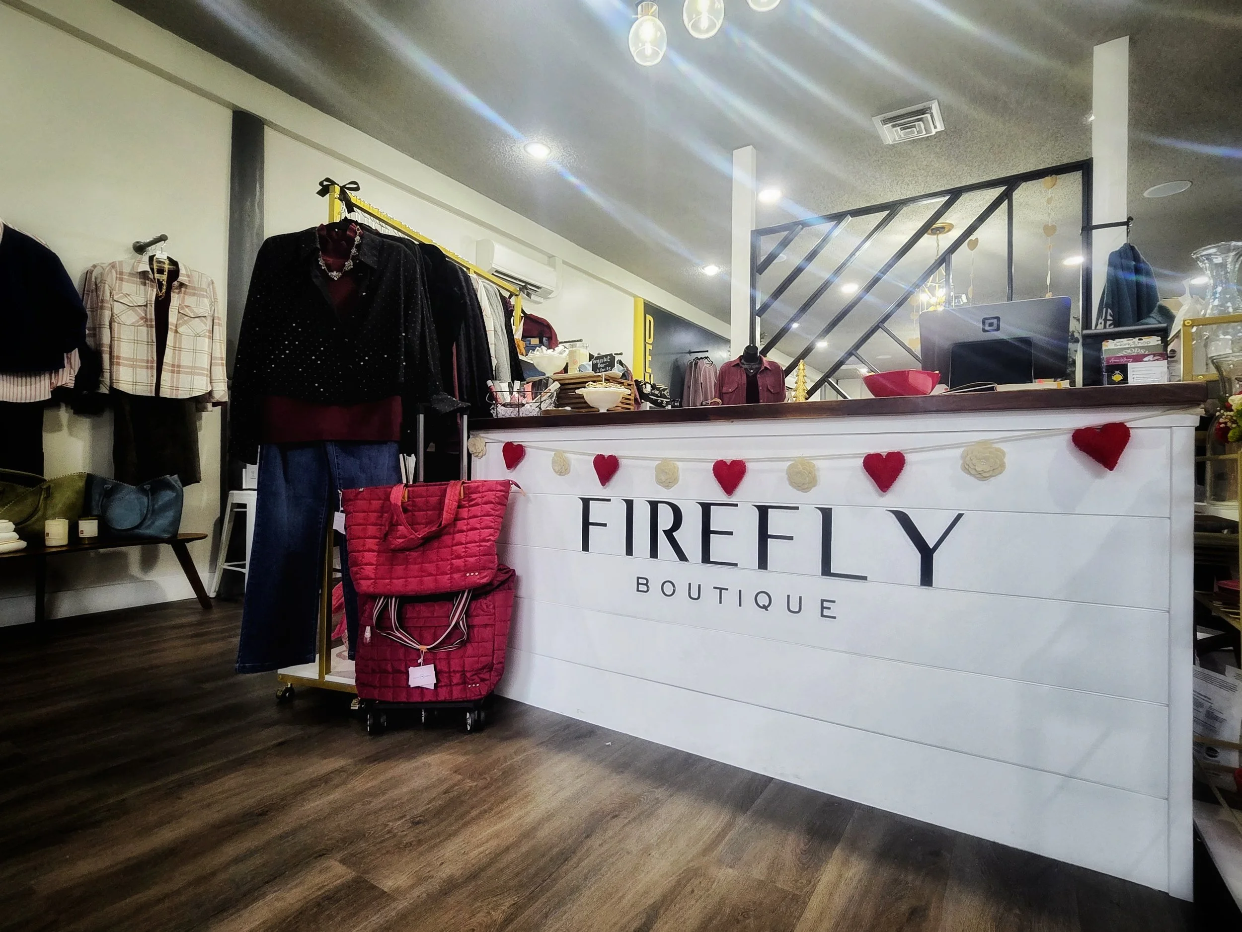 Interior of a boutique named Firefly Boutique with clothing on display, a counter, and pinkHeart and white flower decorations on the counter.