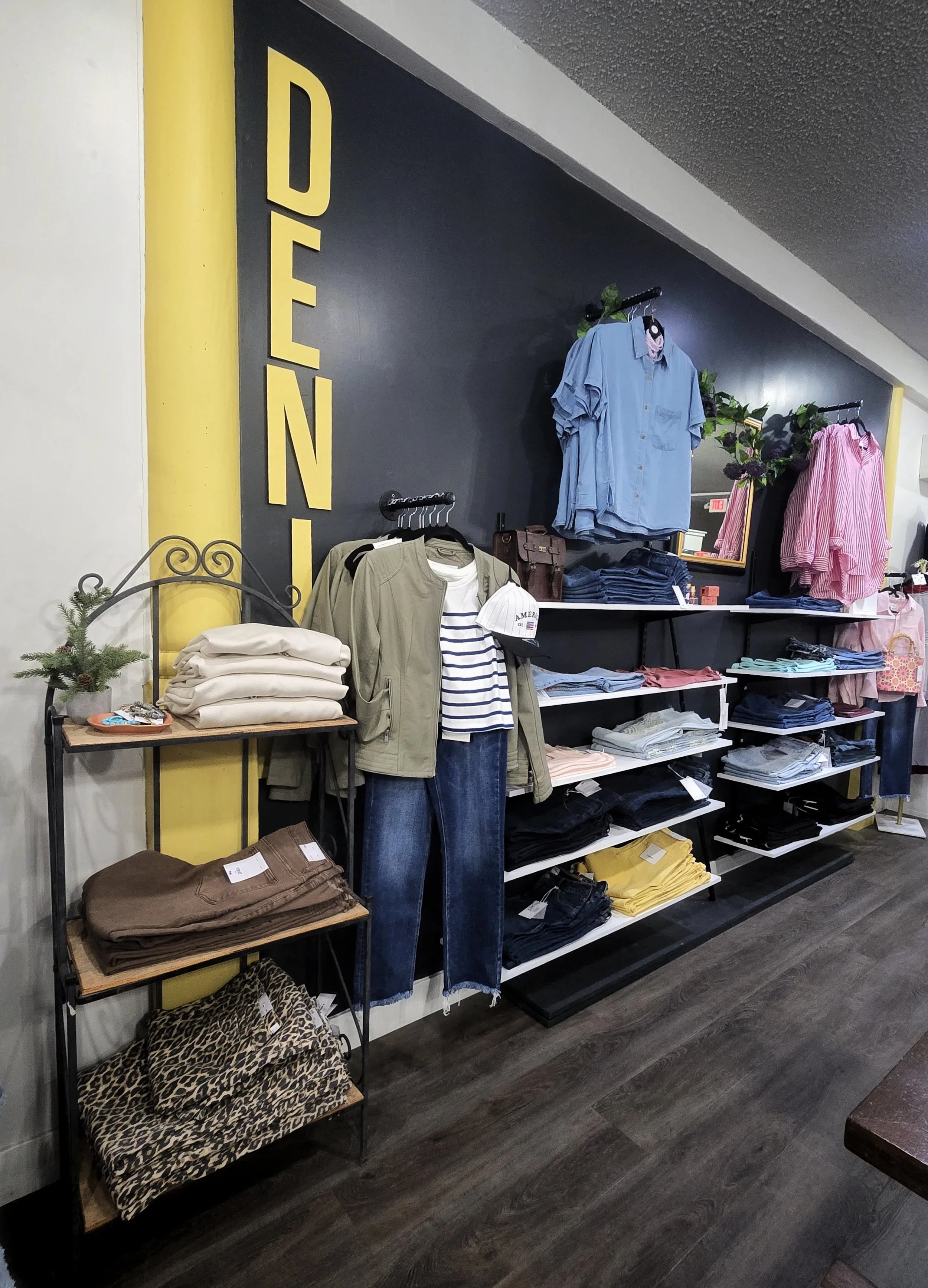 A clothing store display with folded jeans and pants on shelves, hanging shirts, and a small table with folded clothes and accessories.
