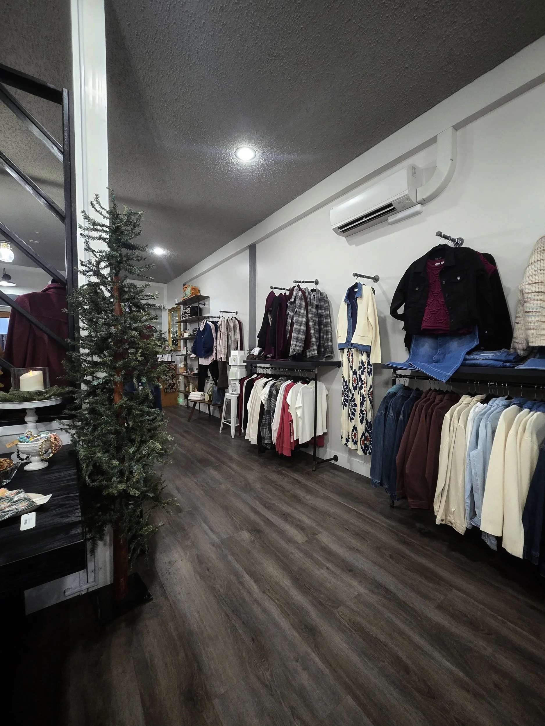 A clothing store interior with racks of jackets, blouses, and pants, a pine tree, and a shelf with various items.