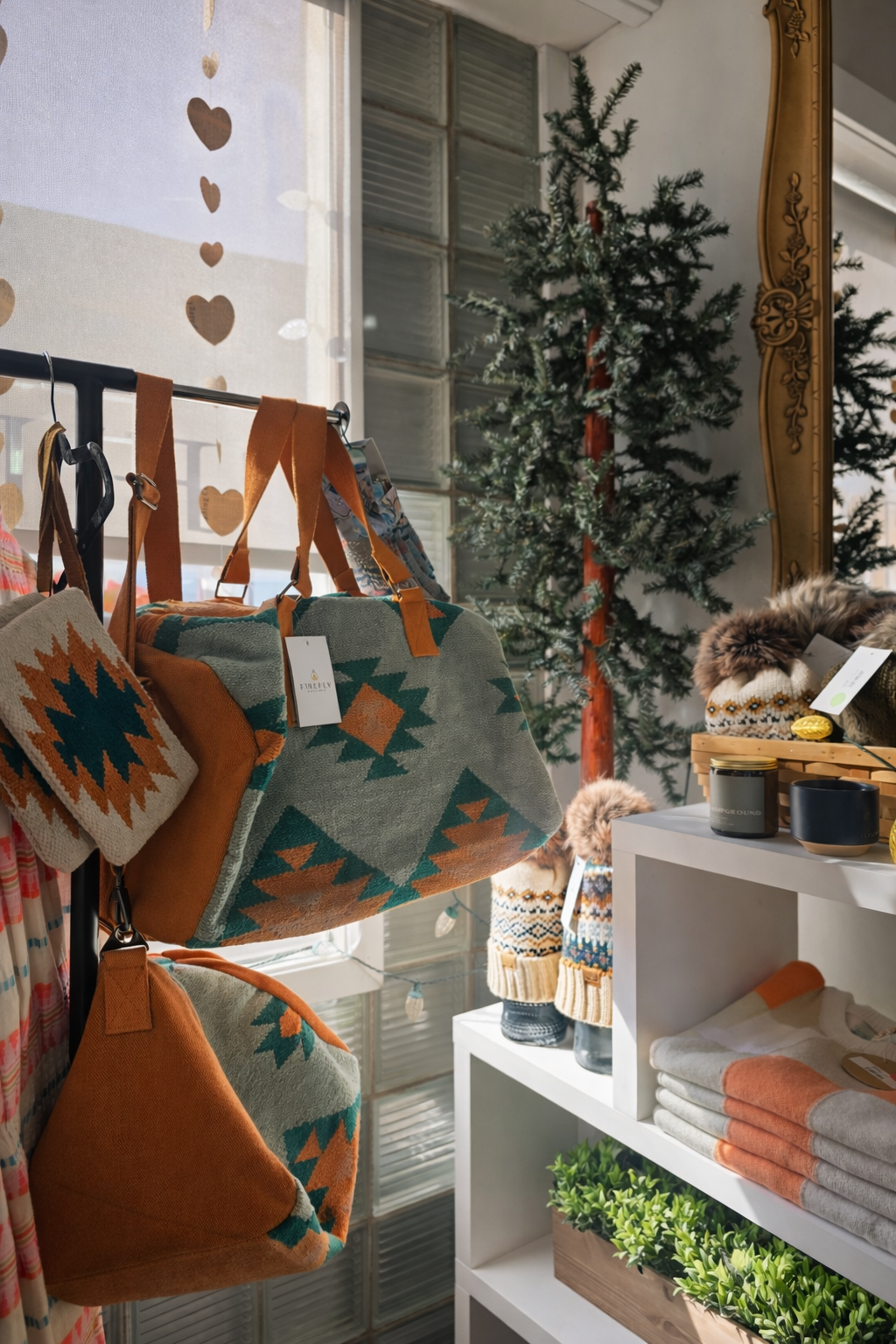 Display of Southwestern patterned handbags, woolen socks, candles, towels, and potted plants inside a store, with a small artificial Christmas tree in the background and sheer curtain with heart pattern on the window.