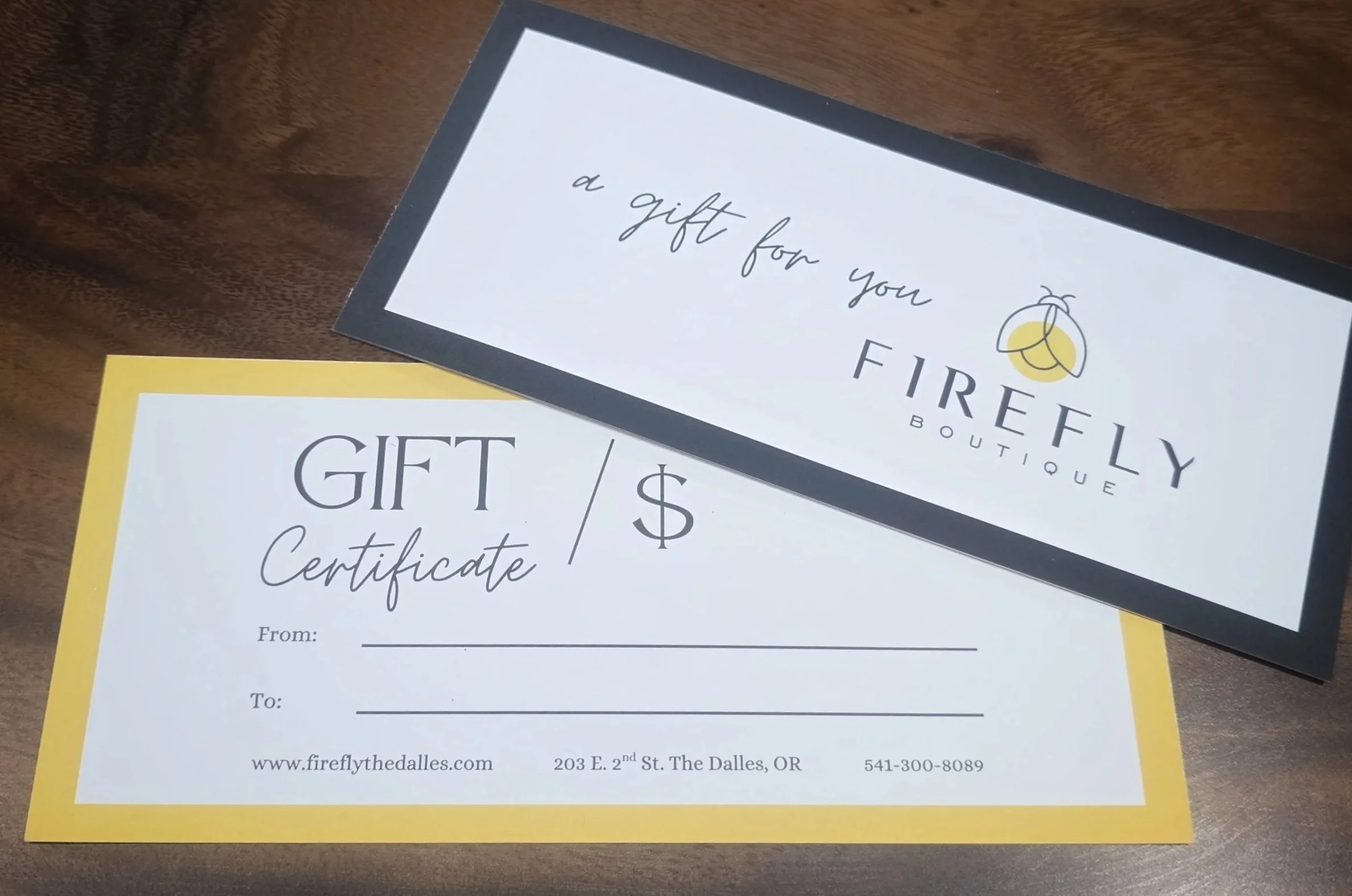 Gift certificate from Firefly Boutique with blank spaces for recipient, giver, and amount, placed on a wooden surface, partially covered by a black-bordered card that says 'a gift for you' from Firefly Boutique with a logo of a bee.