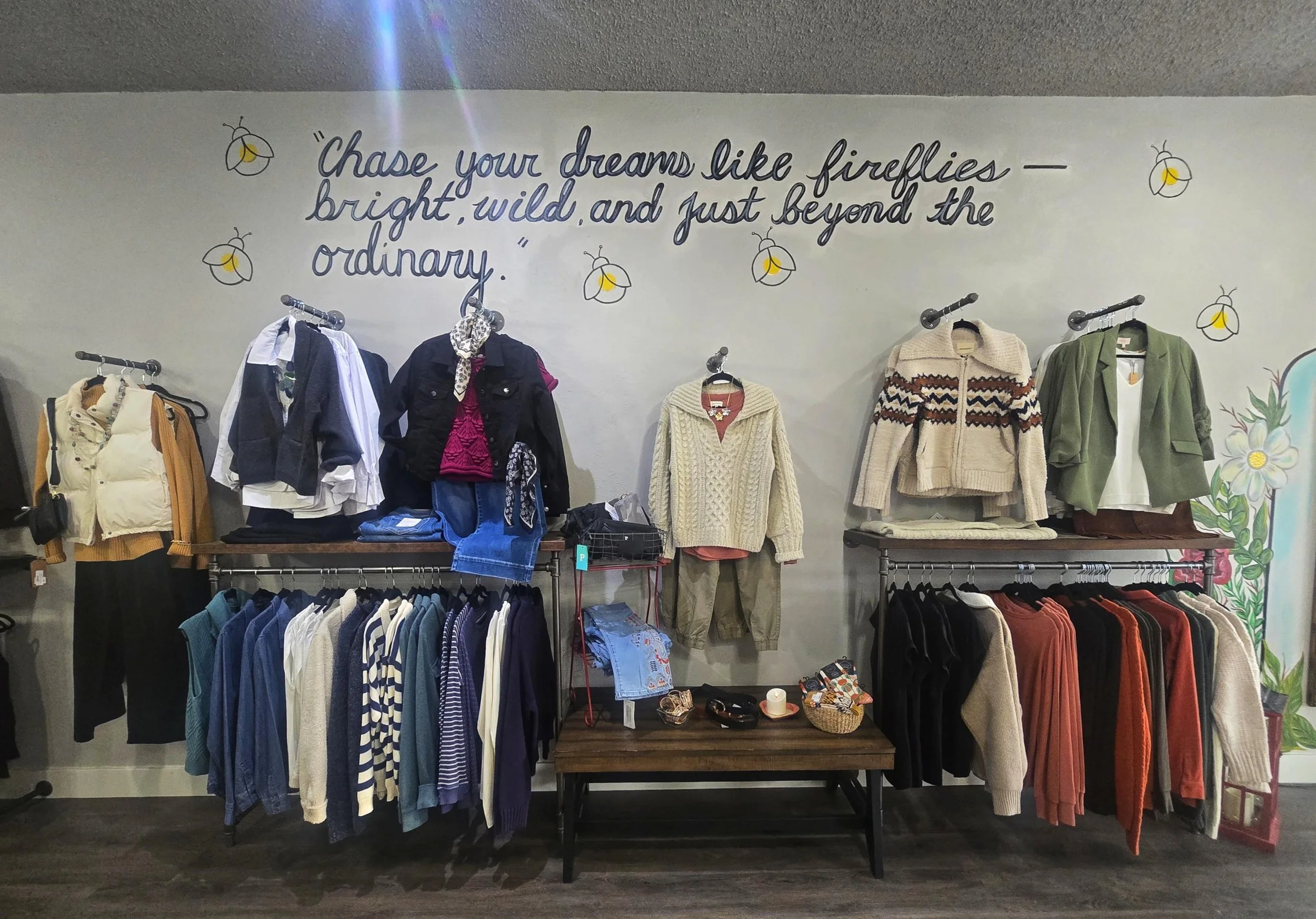 Display of women's clothing on racks and shelves in a boutique store with a quote on the wall that says, 'Chase your dreams like fireflies — bright, wild, and just beyond the ordinary,' surrounded by illustrations of fireflies and flowers.