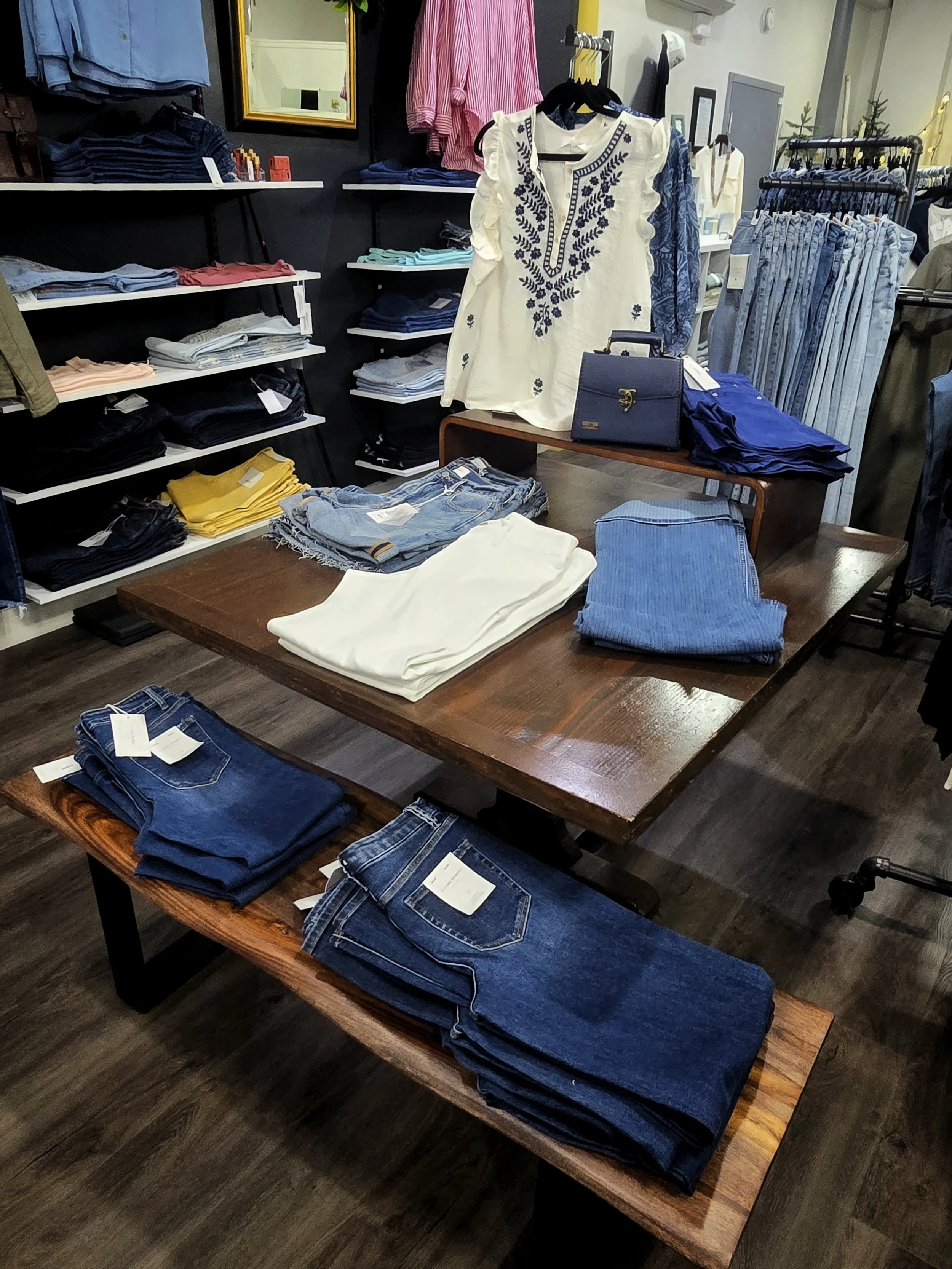 Display of women's jeans and casual clothing in a retail store. Detailing includes folded jeans on tables, a white embroidered top, and various blue and striped clothing items on shelves and racks.