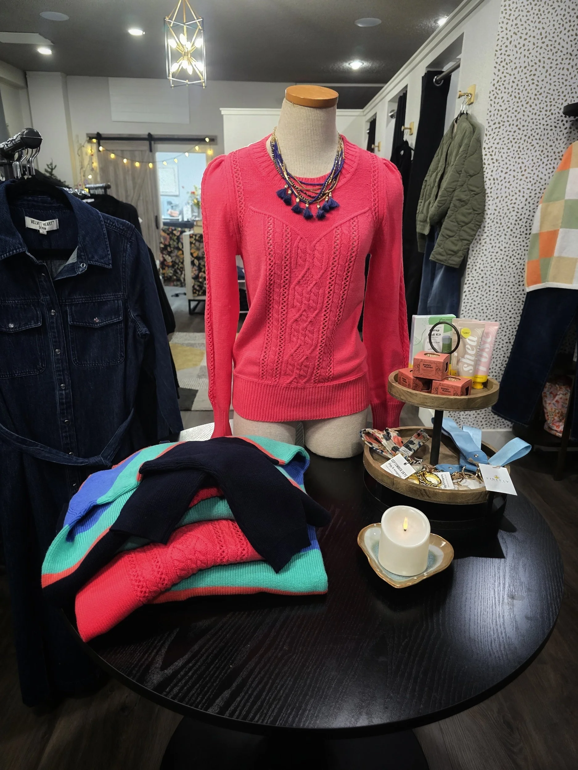 Display of colorful sweaters, jewelry, and accessories on a black round table in a boutique store, with a mannequin wearing a pink sweater and layered necklaces.