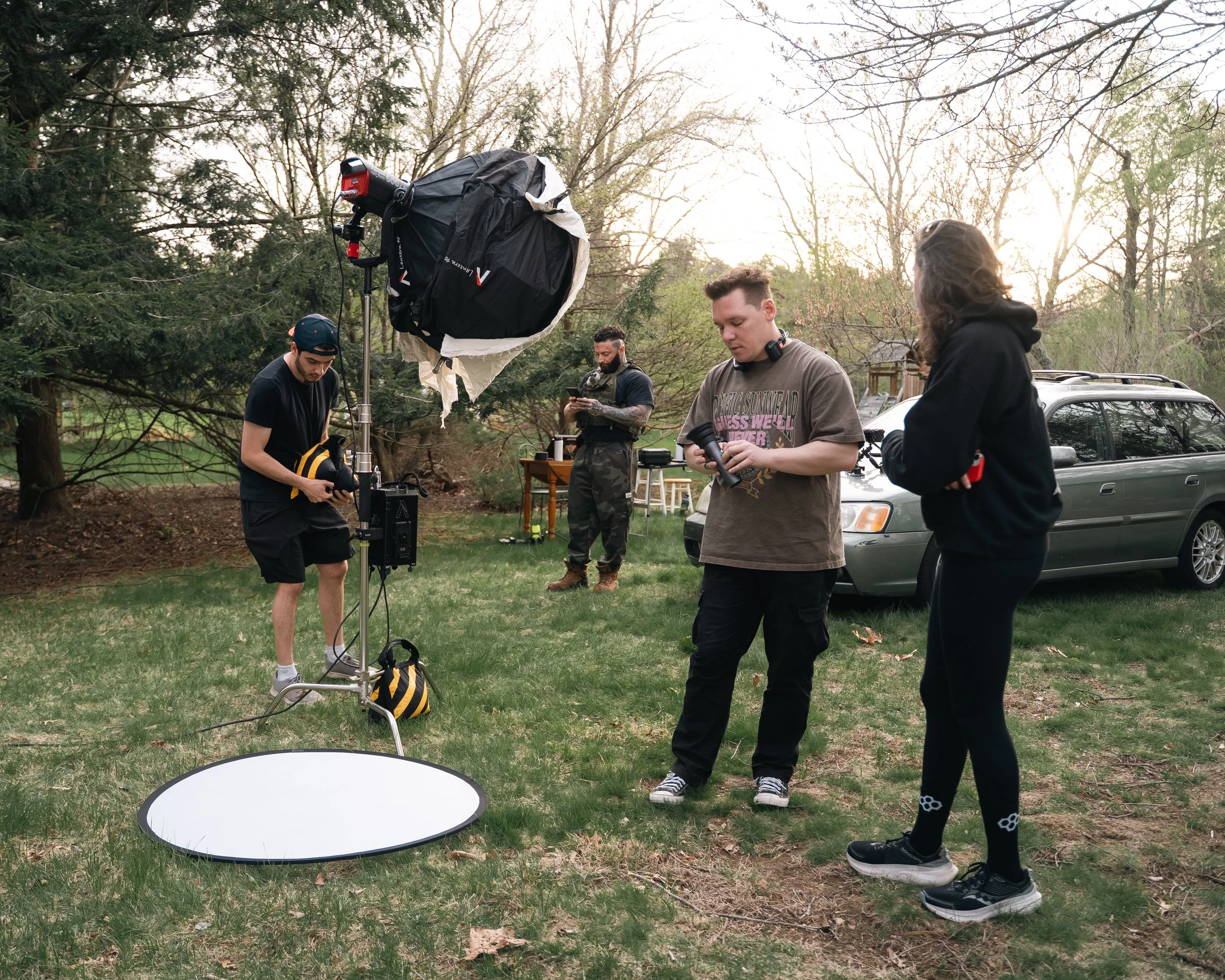 Filming crew setting up equipment outdoors in a park-like area with trees, a car, and a small table in the background during the evening.