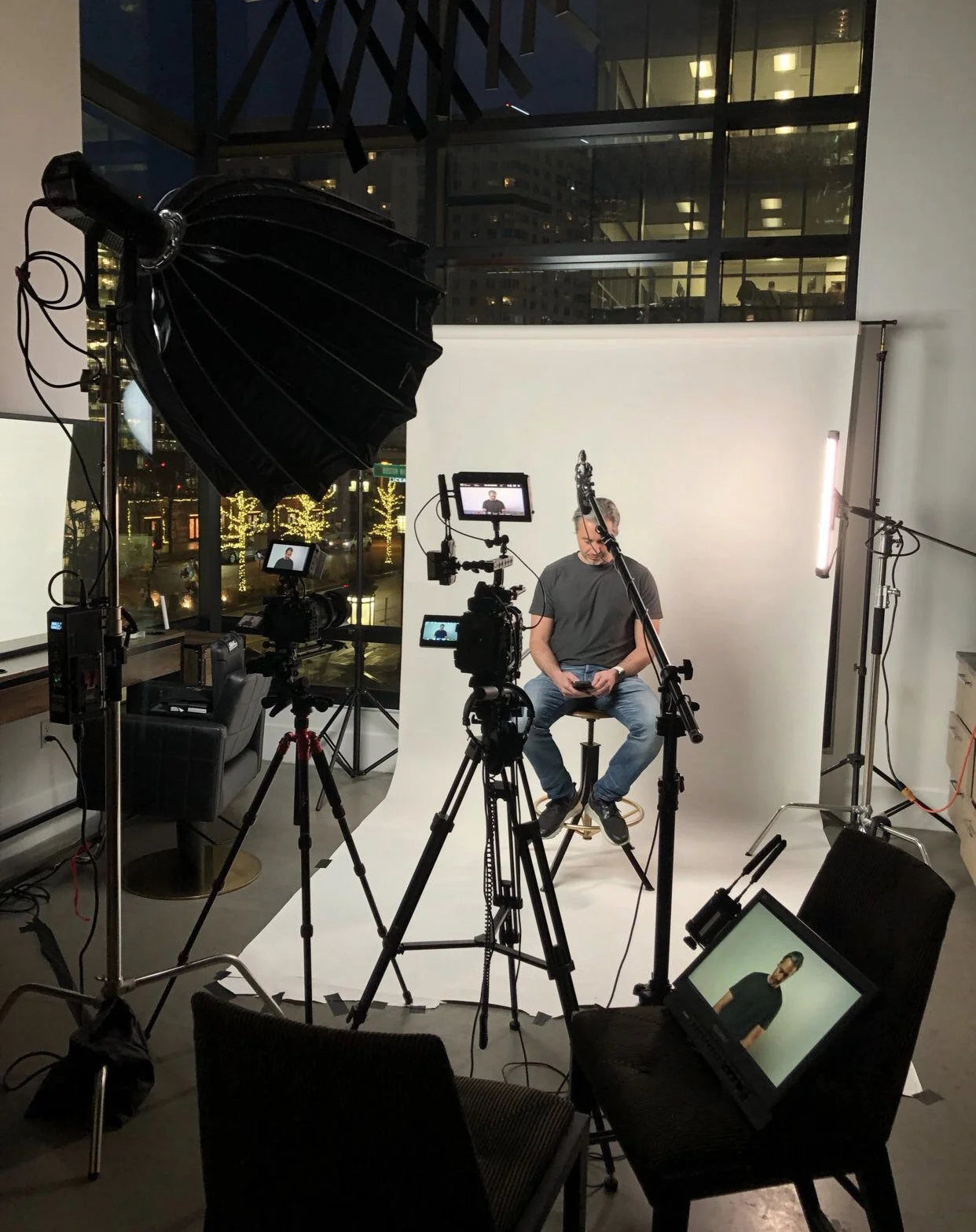 A person sitting on a stool in front of a white backdrop, surrounded by professional photography and video equipment, including cameras on tripods, lights, and monitors, in an indoor studio setting at night.