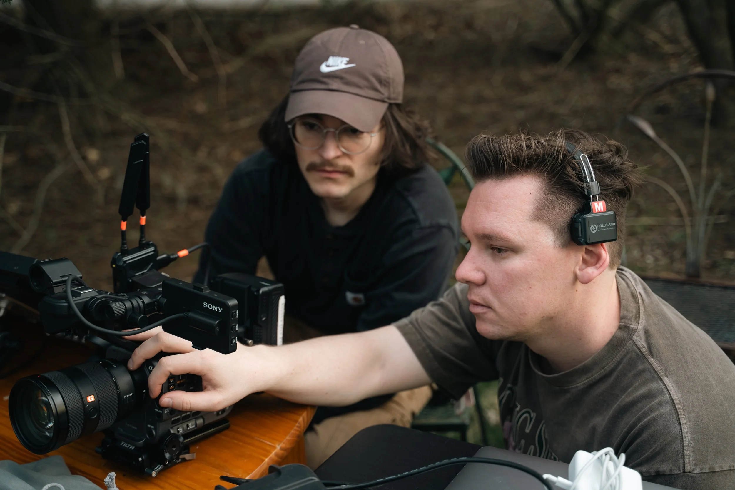 Two men working on a camera setup outdoors the man in front wears a gray t-shirt and headphones, the man behind wears glasses, a brown Nike cap and a black shirt