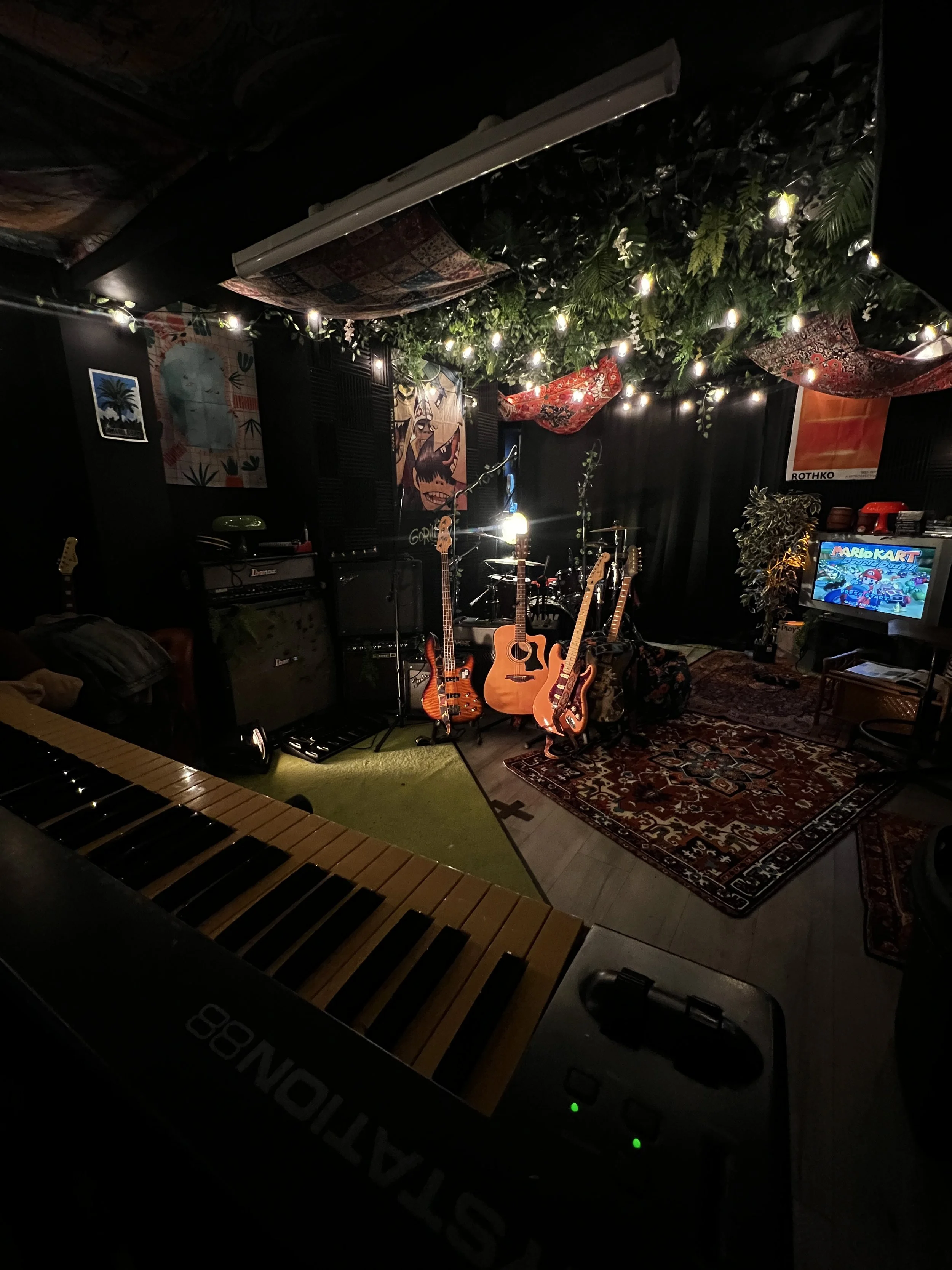 A cozy music studio with string lights and hanging fabric decor. There are multiple guitars, amplifiers, and a drum set. A keyboard is visible in the foreground, and artwork and a TV screen displaying Nintendo Mario Kart are on the walls.