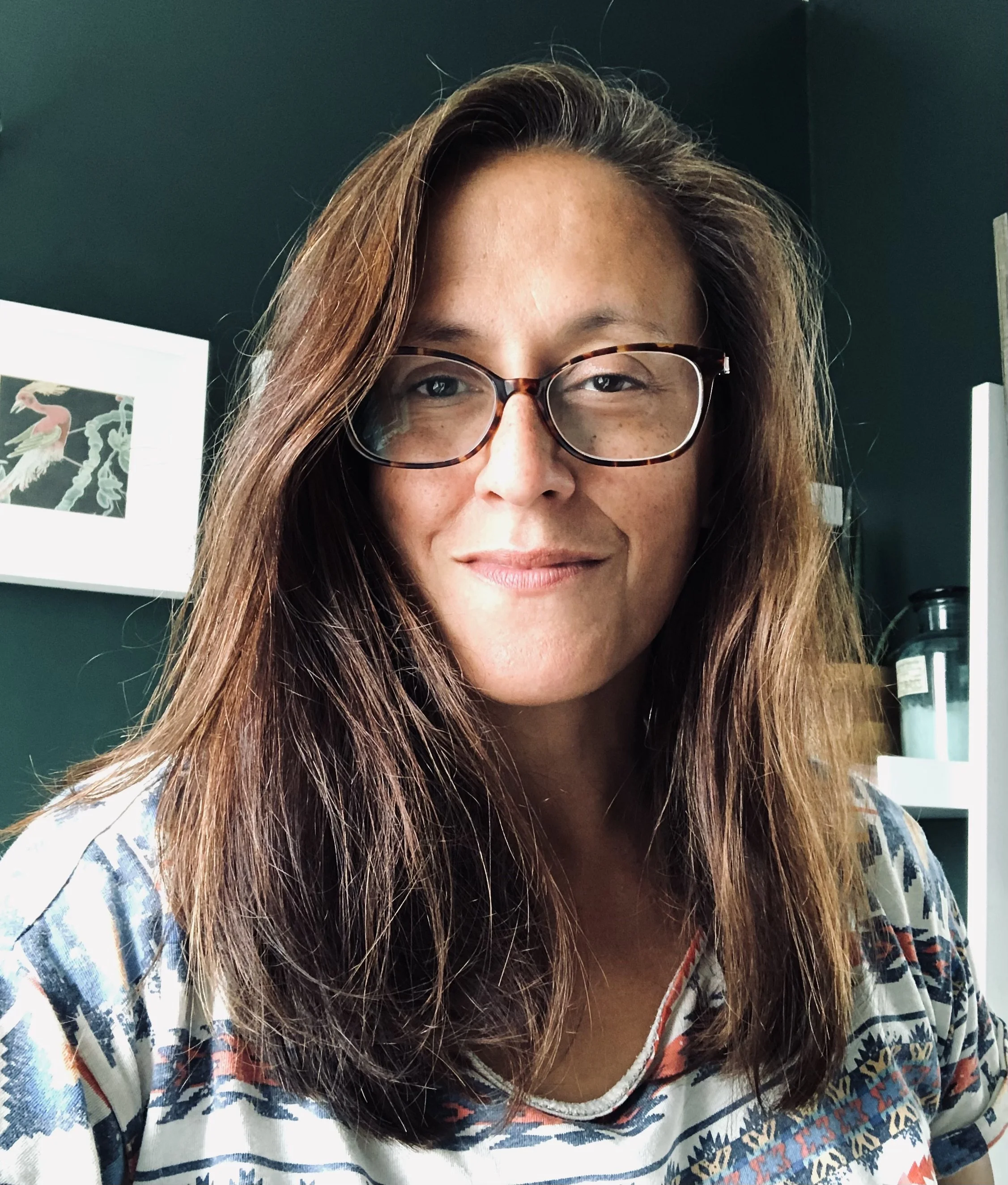 Close-up of a woman with long brown hair and glasses, smiling, indoors with dark green walls and framed artwork in the background.