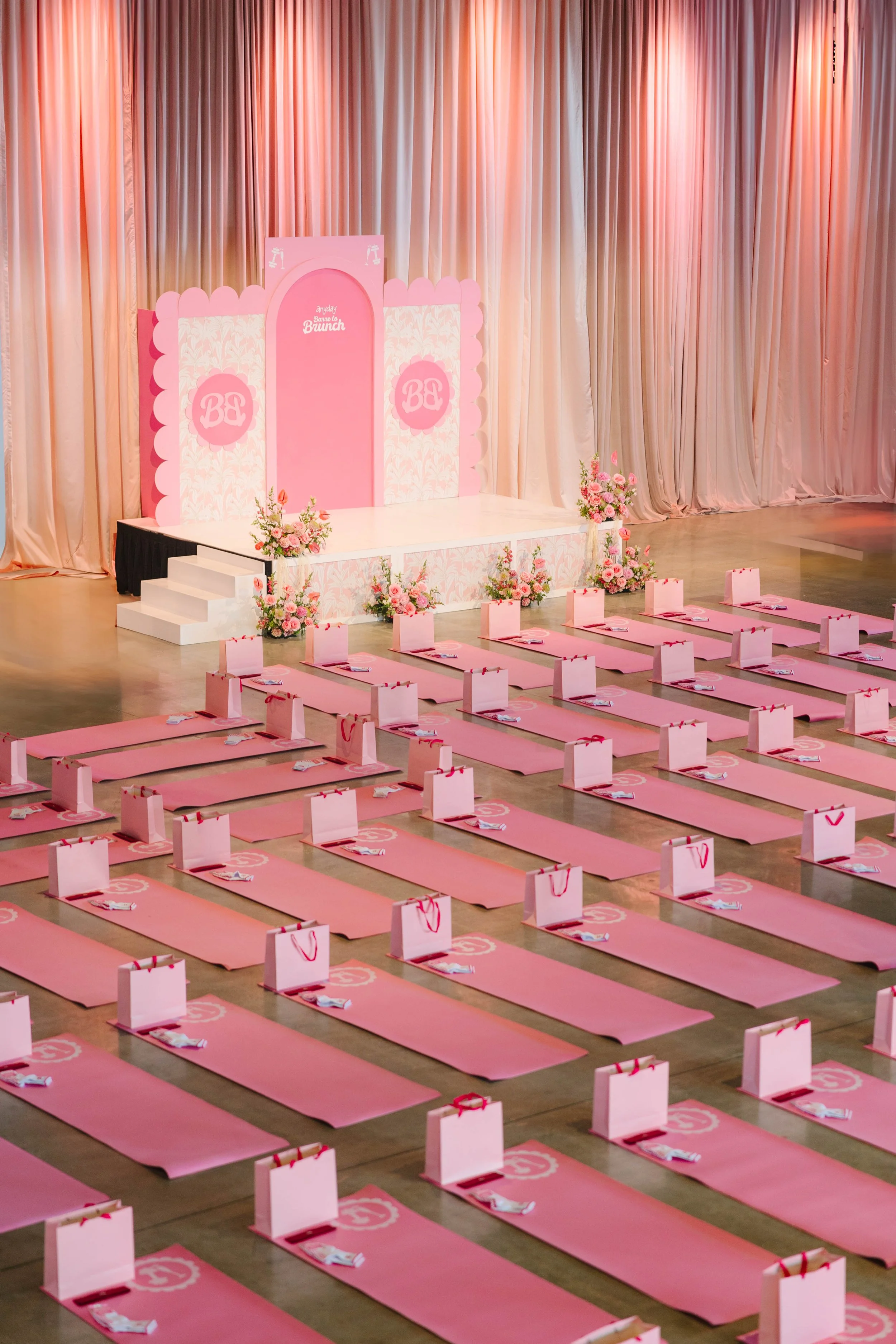 Pink yoga mats with gift bags and rolled towels arranged in rows in front of a decorated stage with pink floral arrangements and curtains, suitable for a girls' brunch event. Barre to Brunch with Jenna Palek