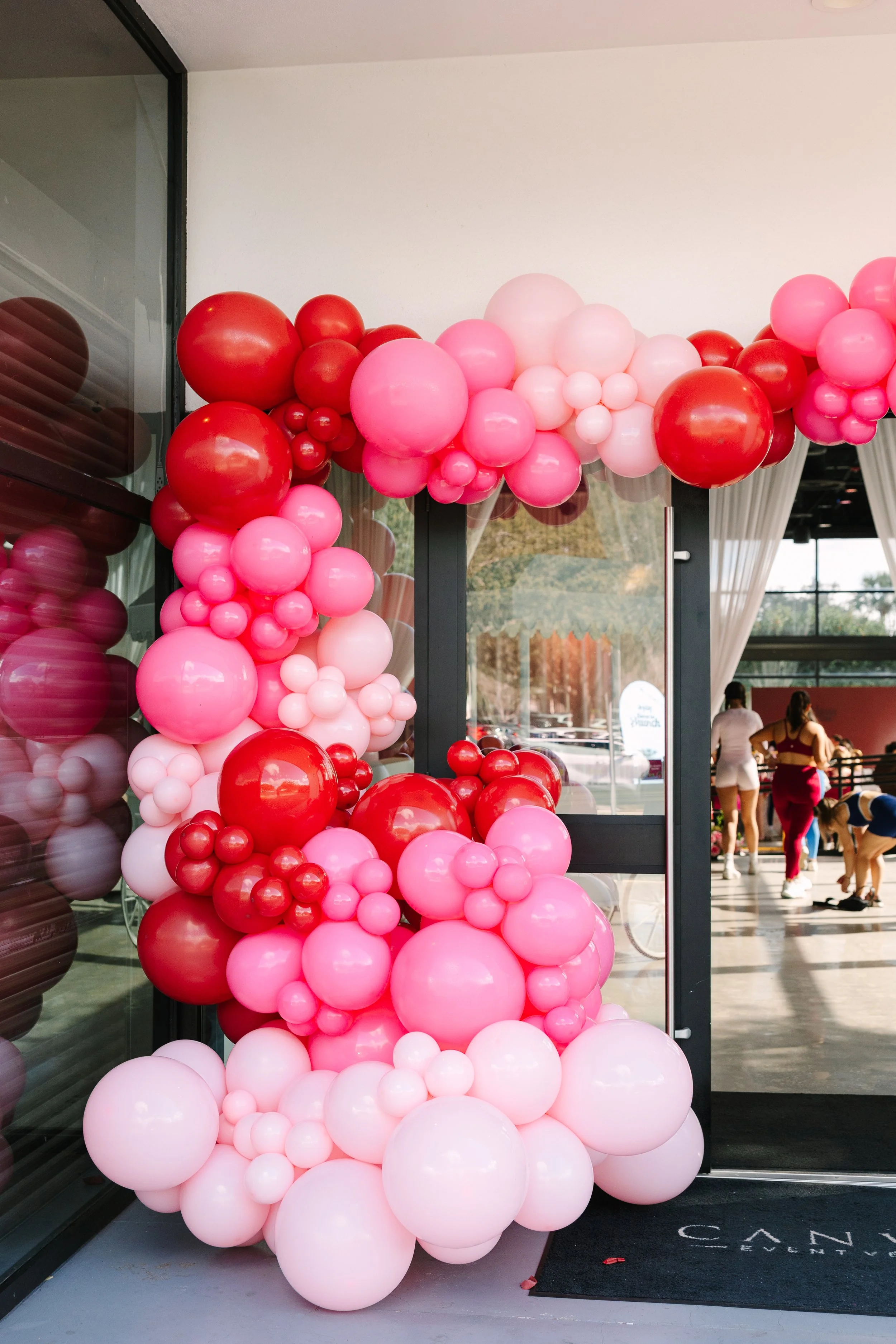 Decorative balloon arrangement with pink, red, and white balloons forming an arch and cluster at the entrance of a building. Barre to Brunch with Jenna Palek